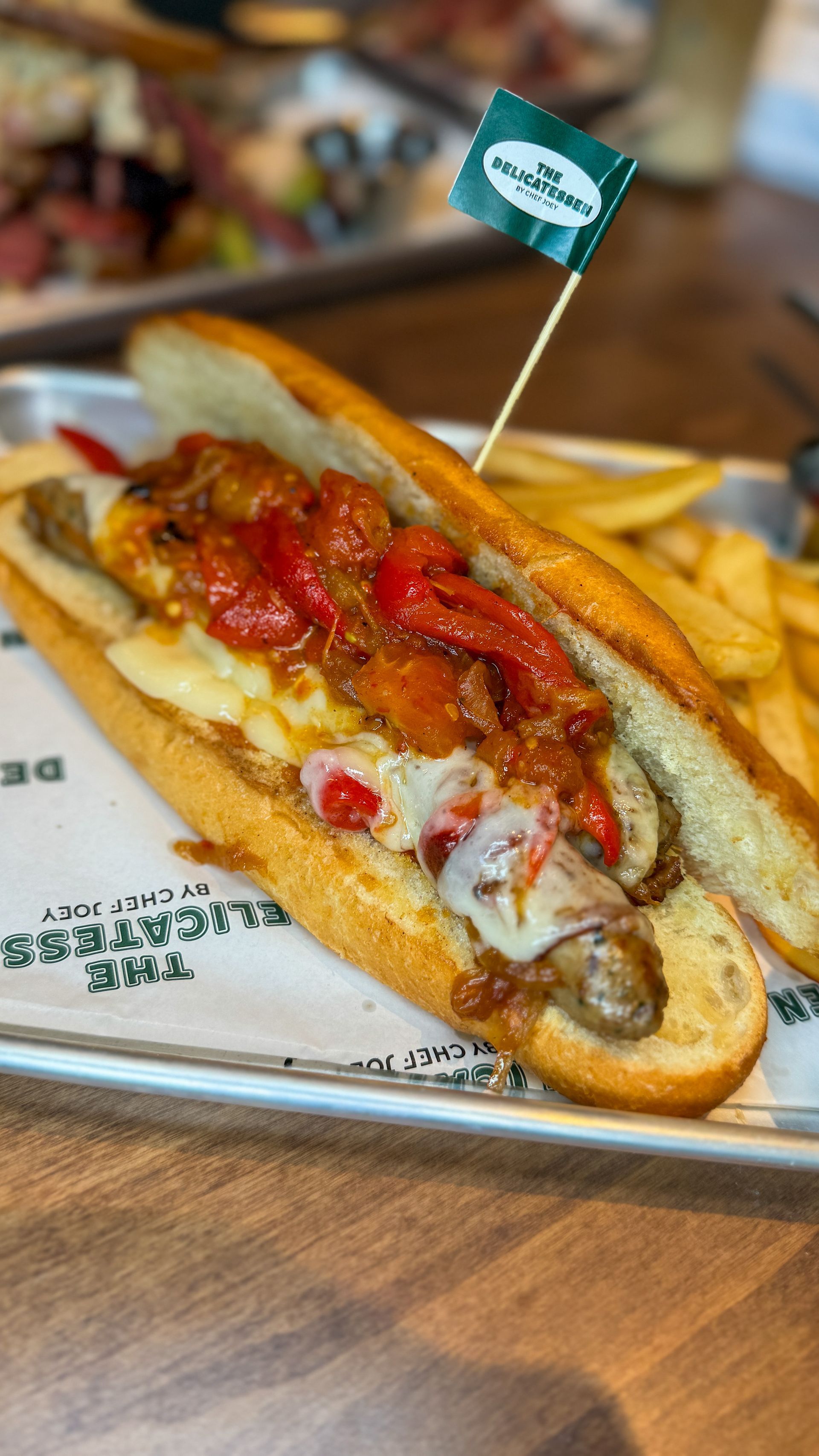 Sub sandwich with sausage, peppers, cheese, and fries on a tray. A green flag is inserted into the sandwich.