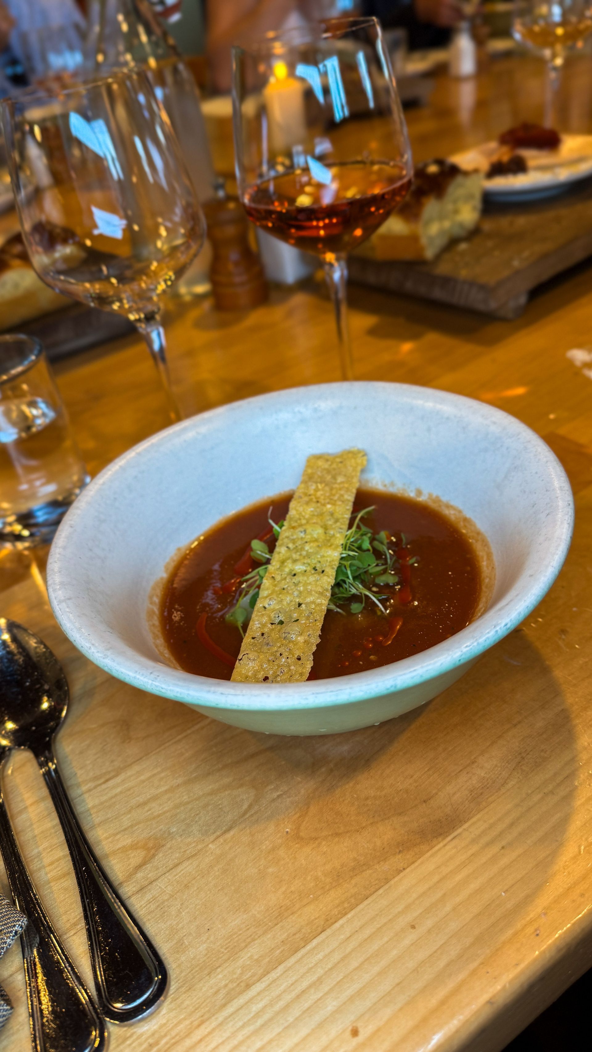 White bowl of dark soup garnished with herb and crisp bread on a wooden restaurant table, with wine glasses behind