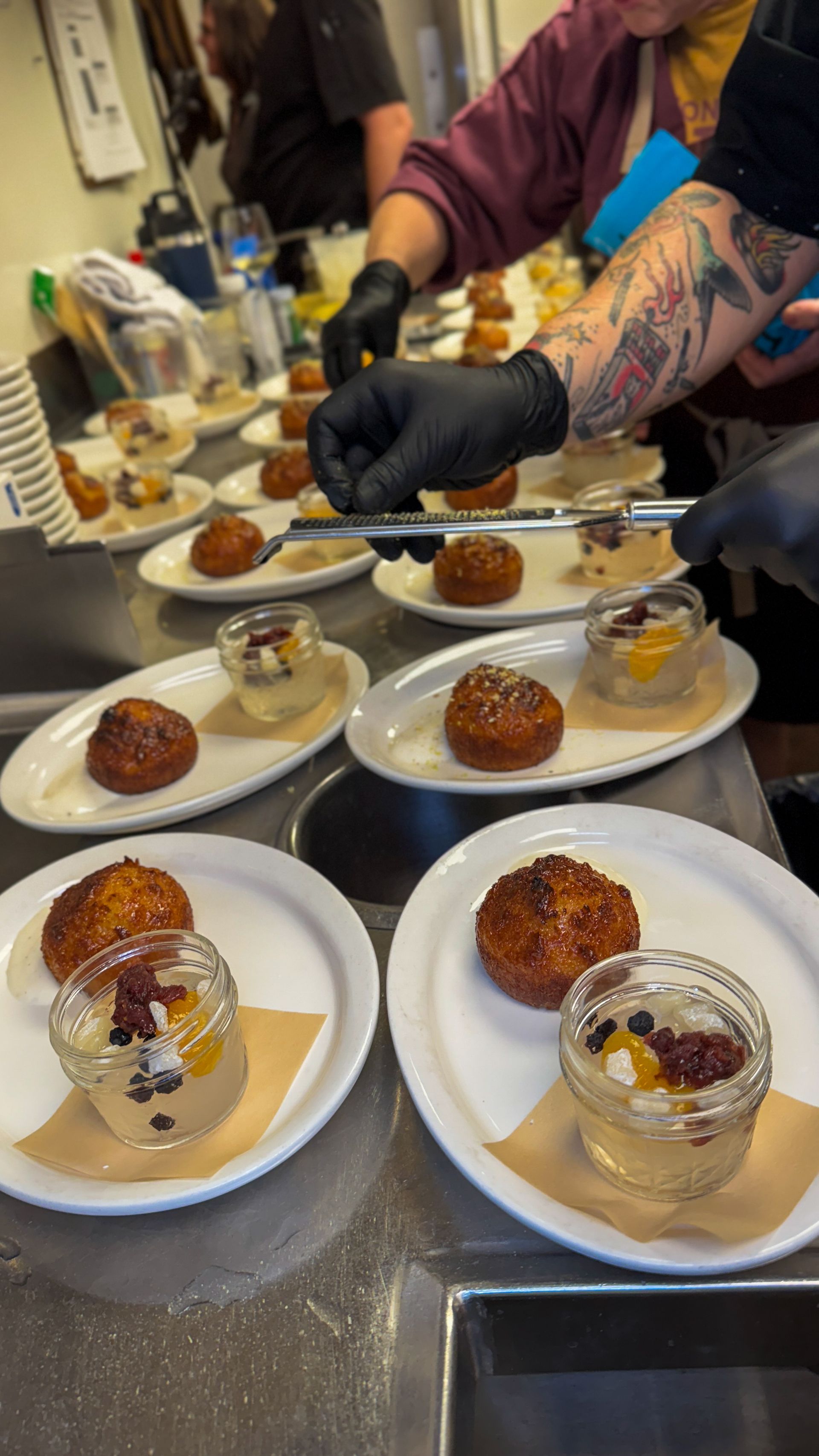 Desserts being plated by a tattooed worker in black gloves at a kitchen prep counter.