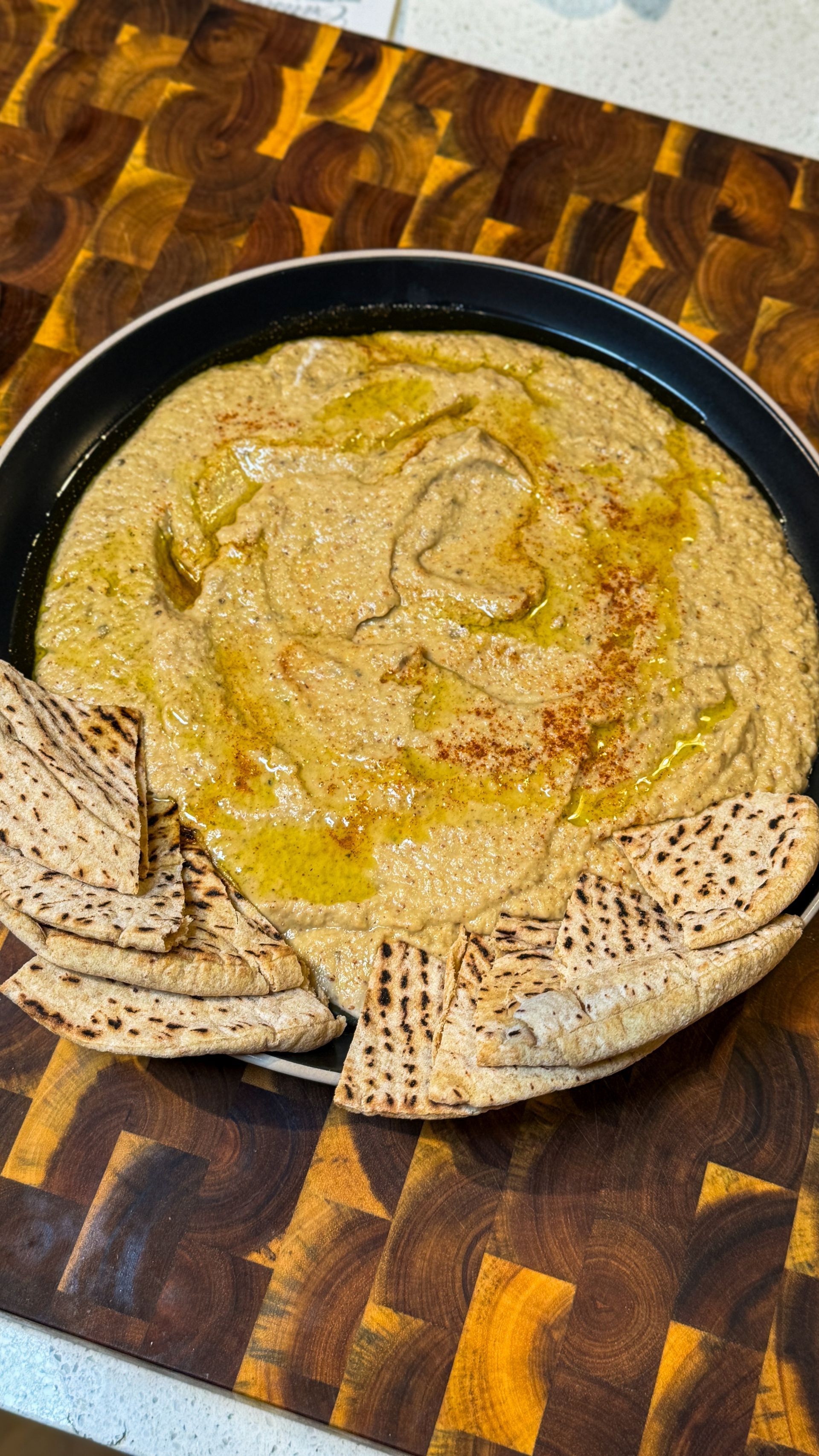 Hummus with pita bread in a pan, drizzled with oil and paprika, on a wooden cutting board.
