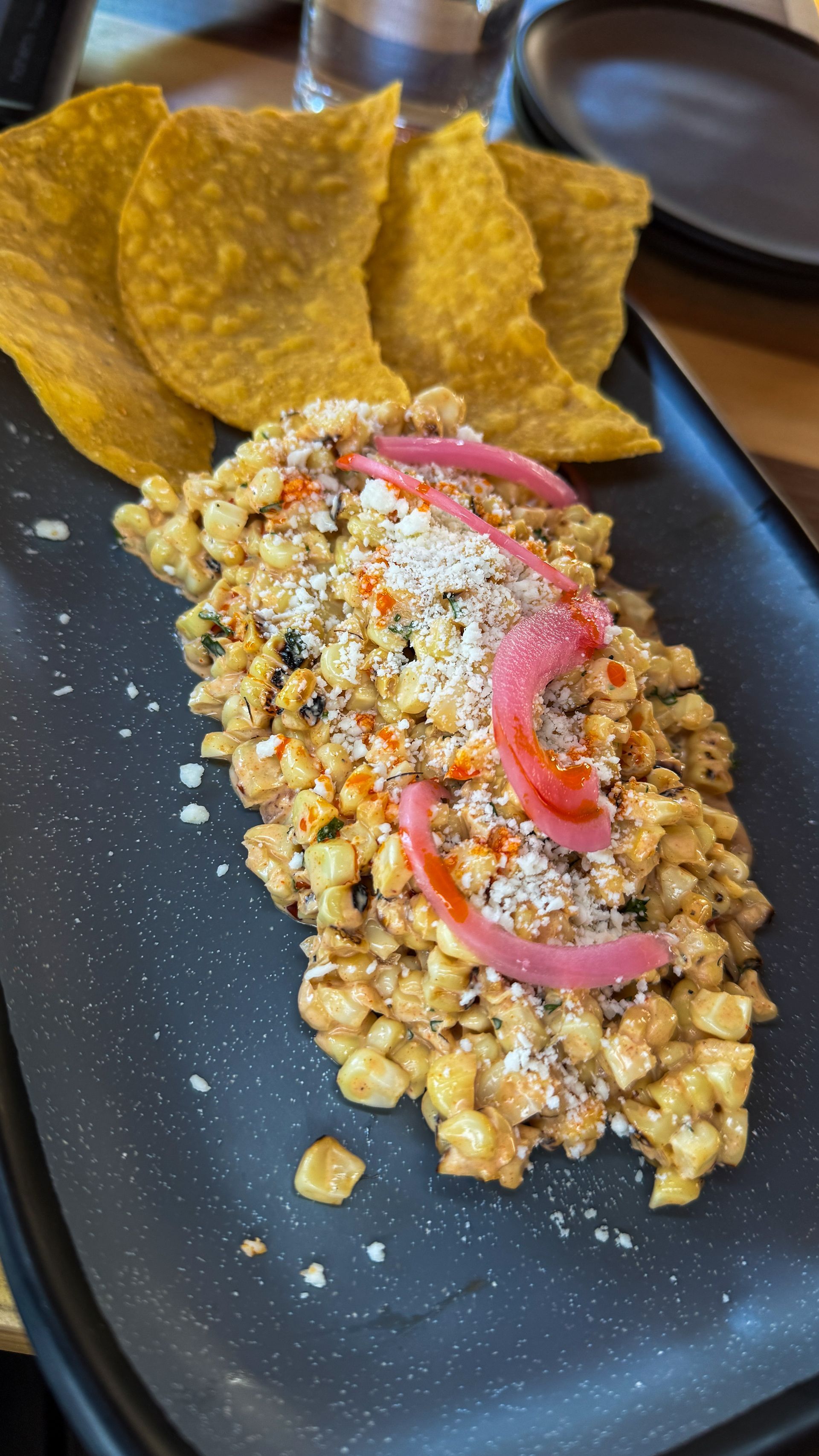Crunchy chips beside a golden corn salad topped with pickled red onions on a black plate.