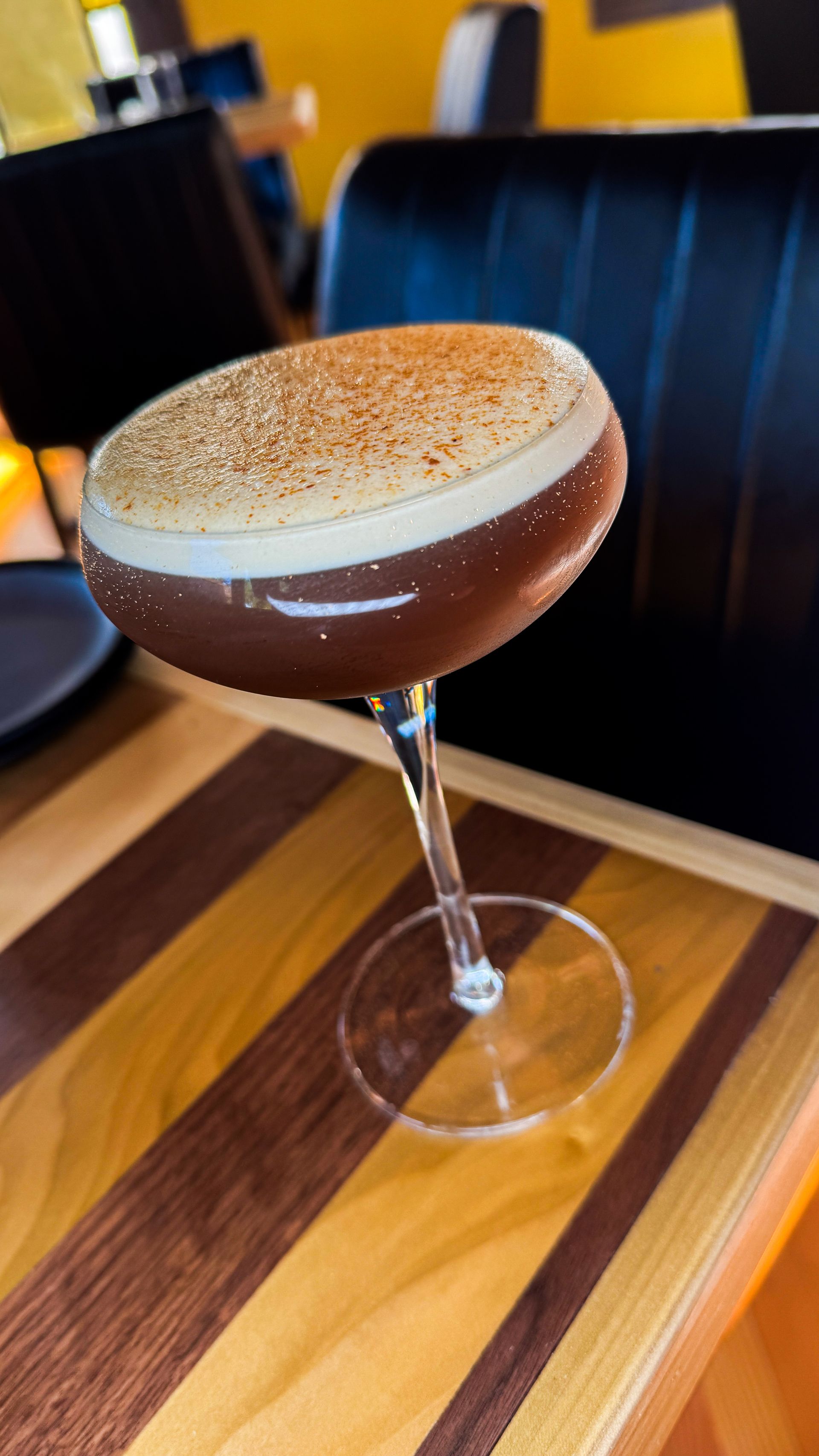 Chocolate martini in a coupe glass on a striped wooden bar top, topped with frothy foam and cocoa powder.