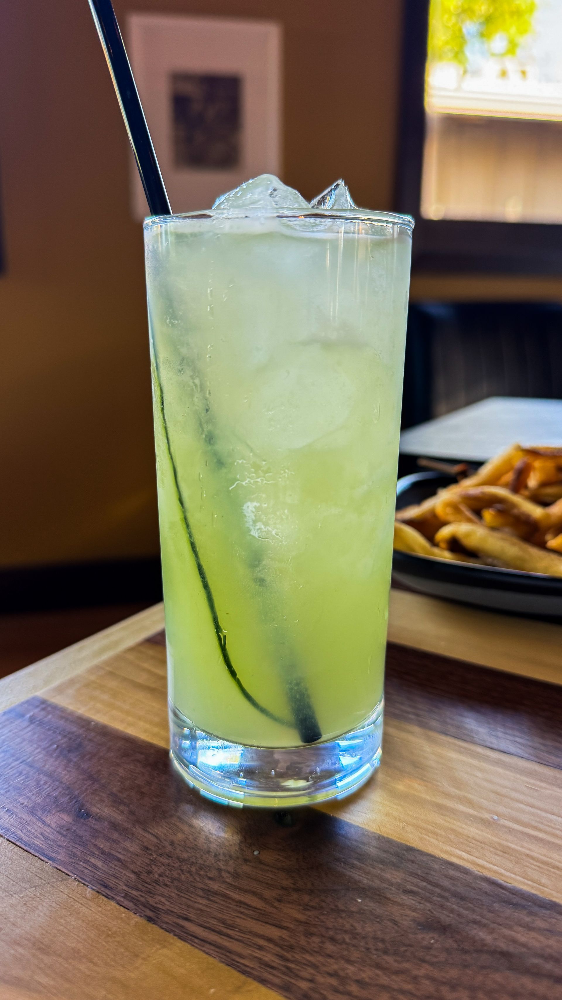 Tall glass of pale green iced drink with a black straw on a wooden table, with fries in the background
