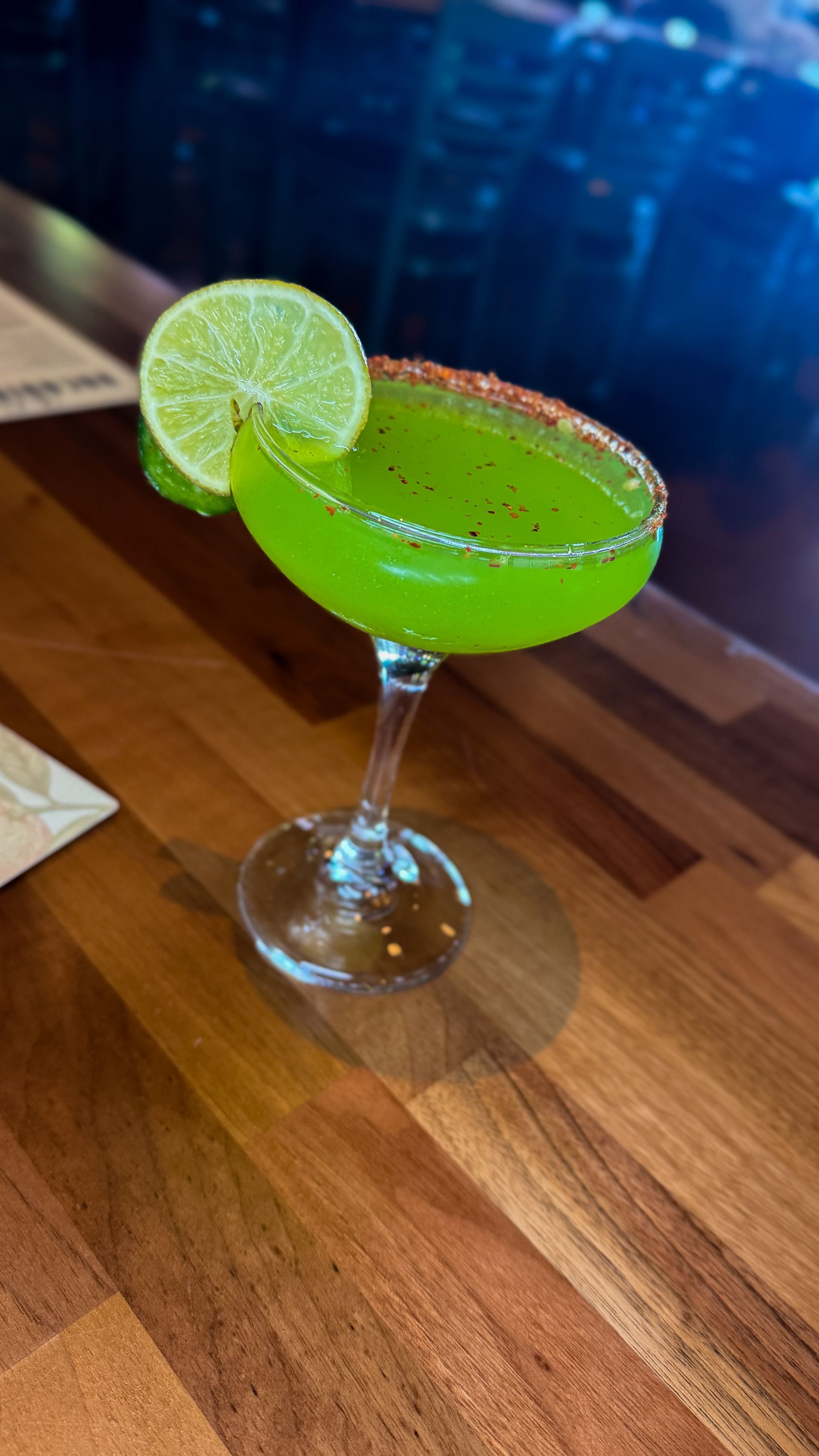 Green cocktail in a stemmed glass with a lime garnish and a spicy rim, on a wooden surface.