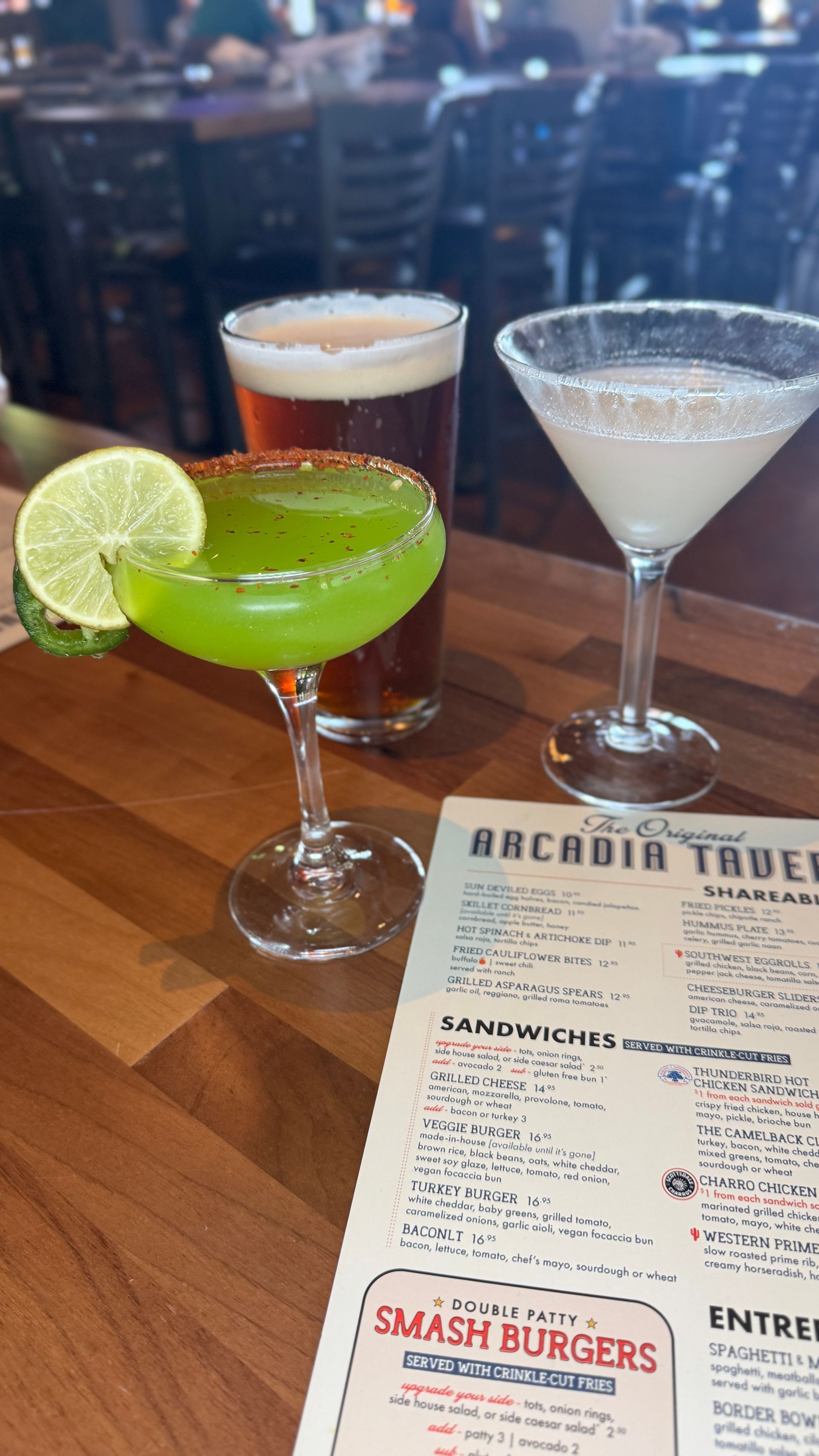 Three cocktails and a beer on a wooden table with a menu in a bar setting.