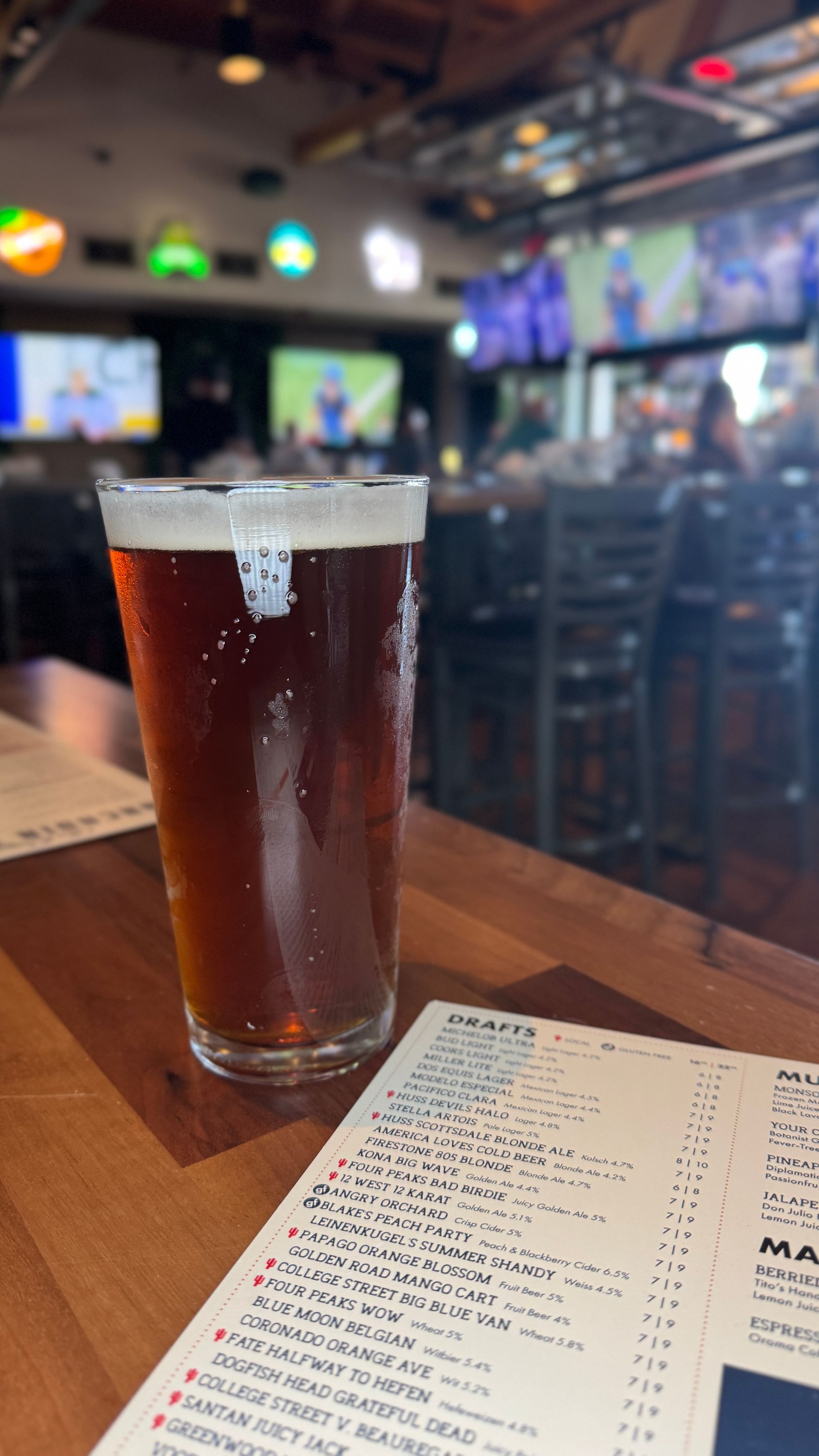 A pint of amber beer on a wooden table, next to a menu, in a sports bar.