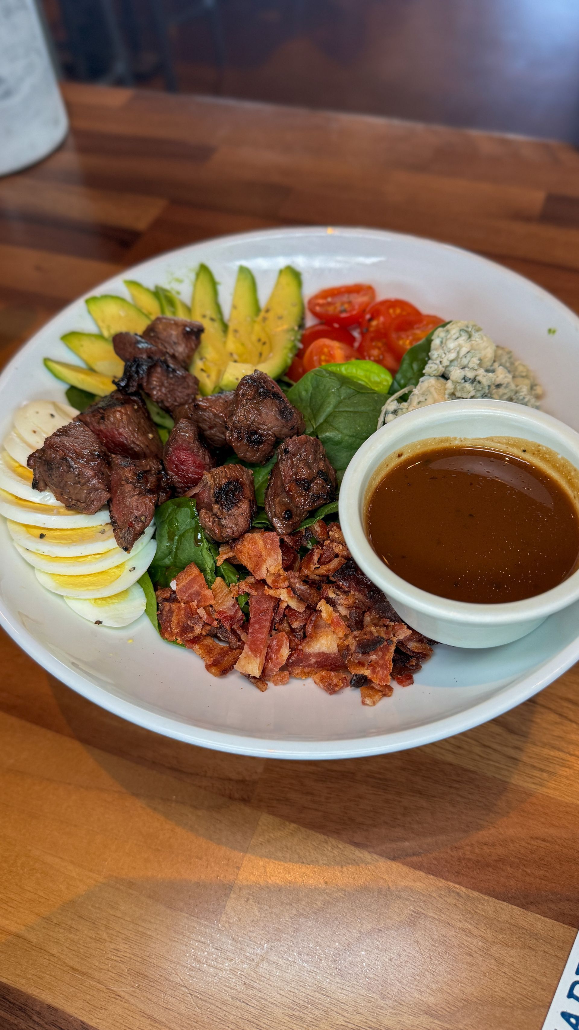 Salad bowl with steak, avocado, eggs, bacon, tomatoes, greens, quinoa, and dressing.