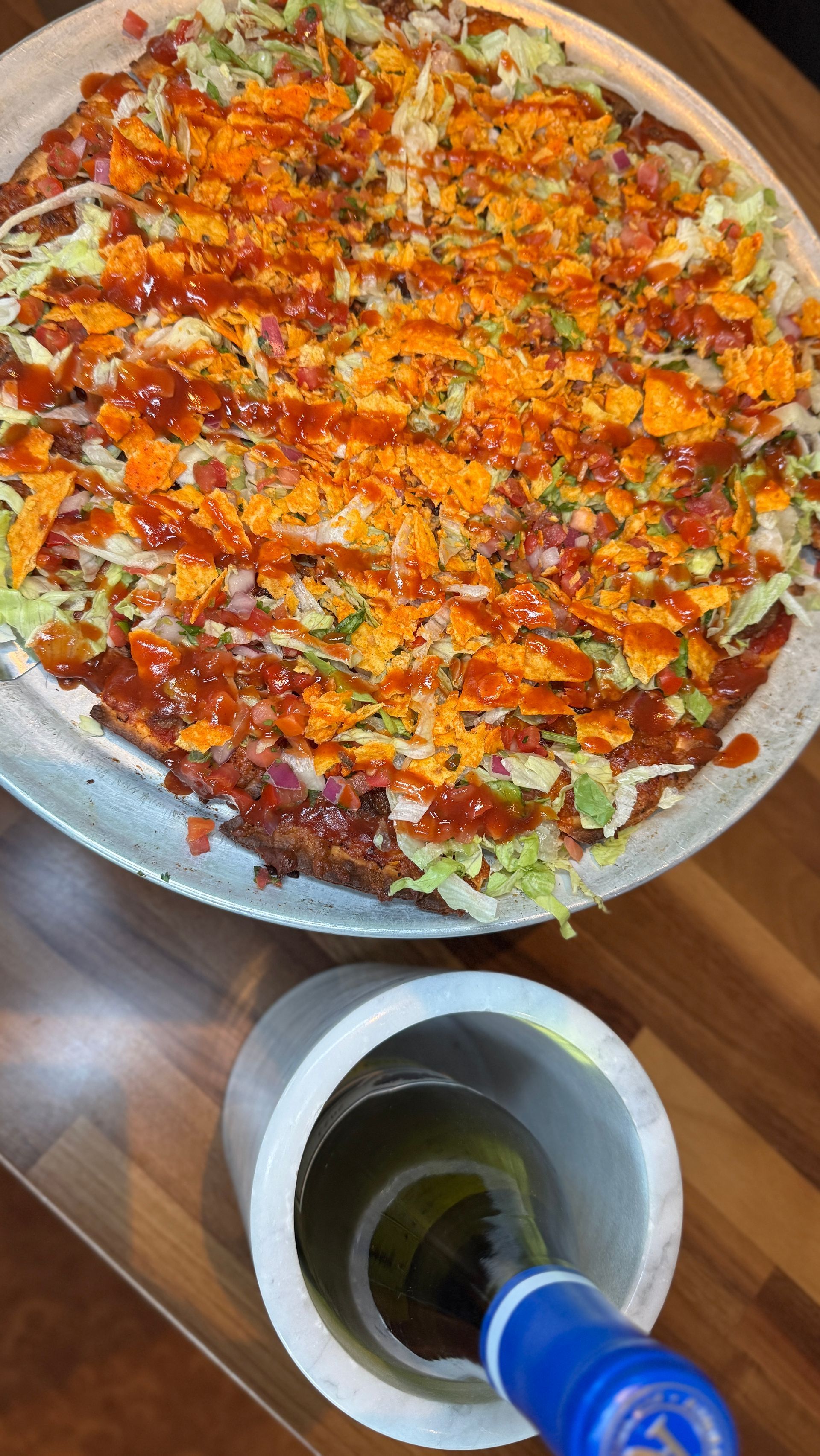 Pizza with toppings on a metal tray next to a bottle of white wine in an ice bucket on a wooden table.