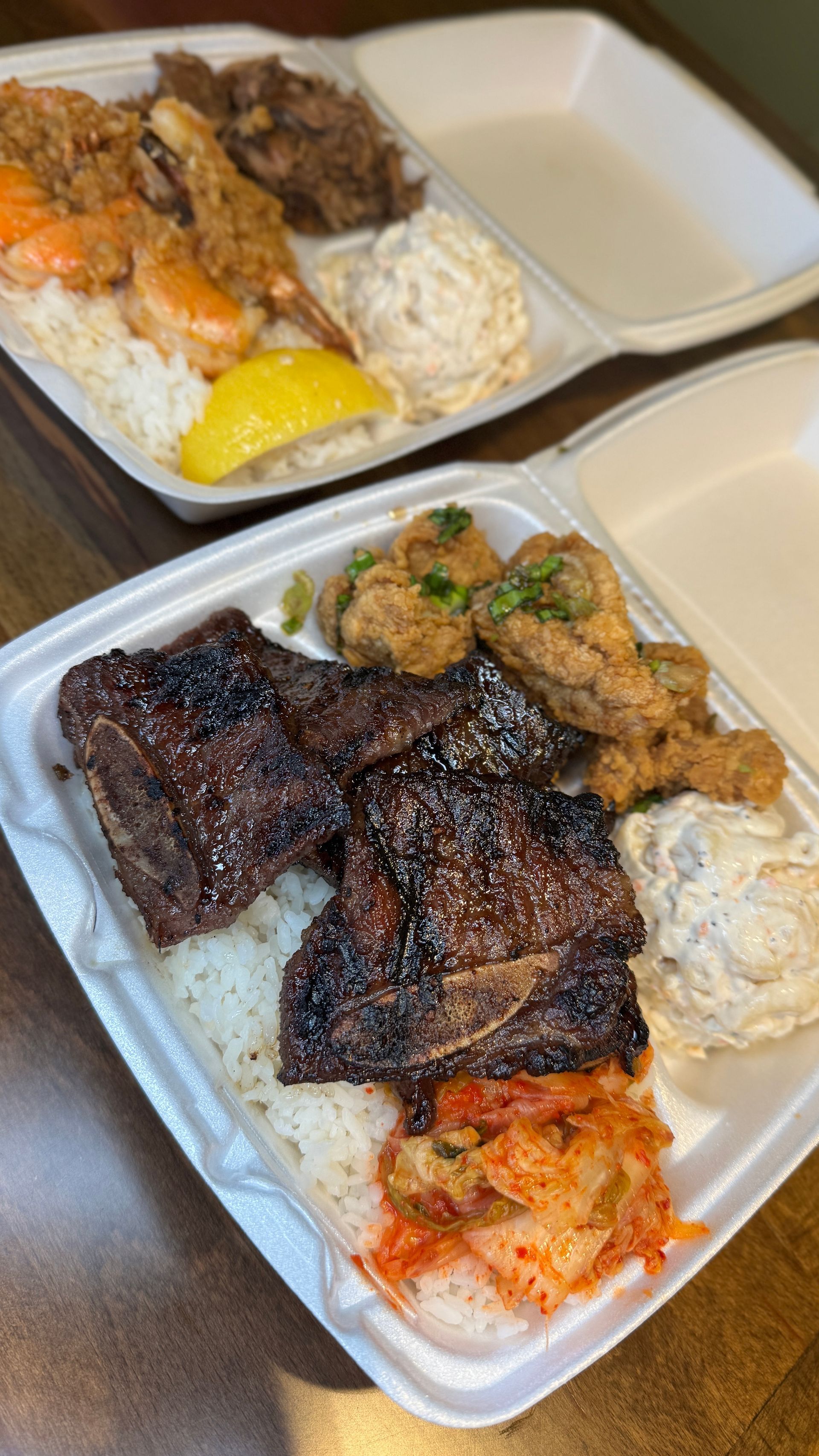 Two takeout boxes with rice and grilled meat, topped with sauce and a yellow side dish