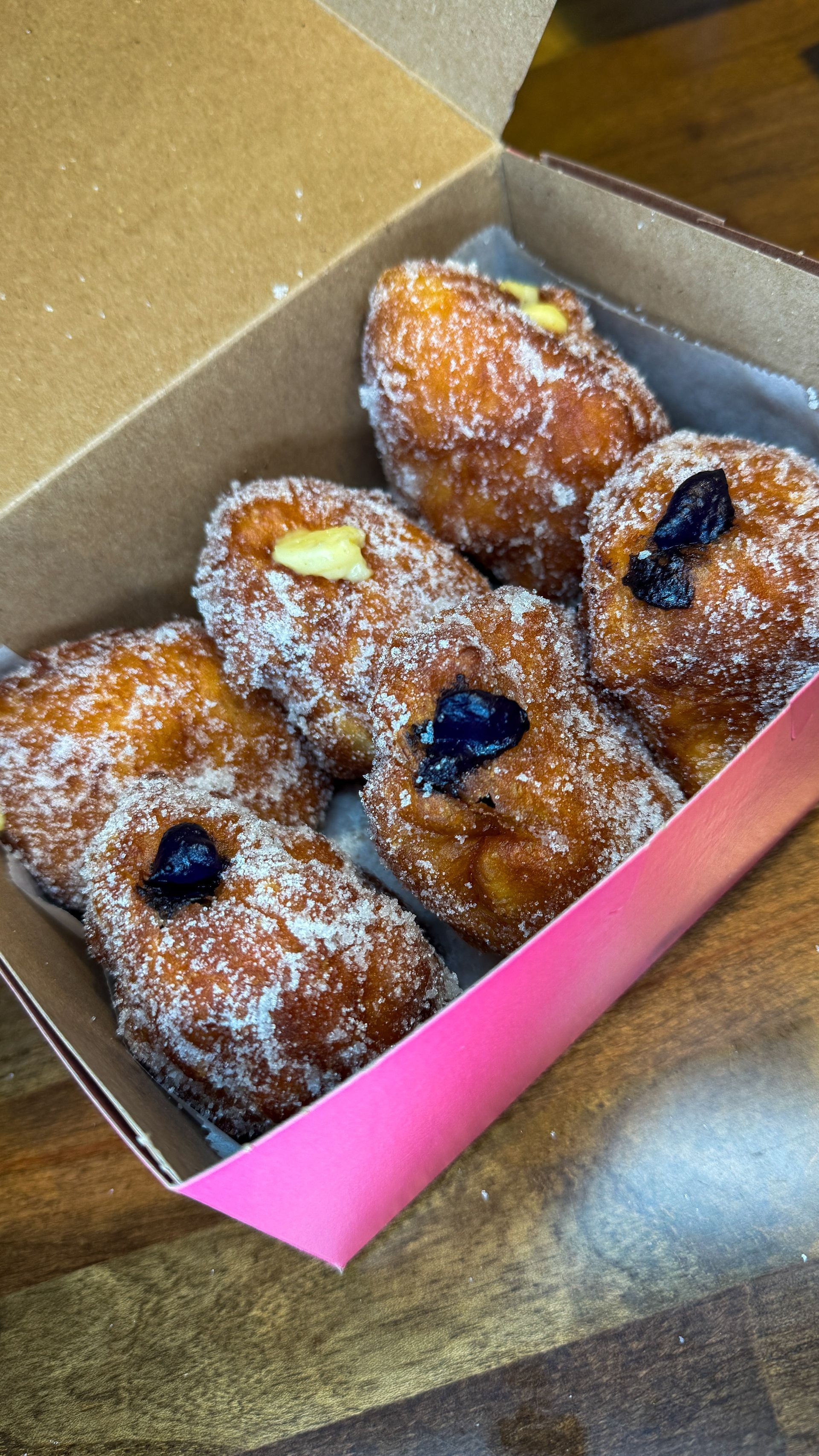 Box of sugar-coated donuts, some filled with cream and topped with berry compote, on a wooden surface.