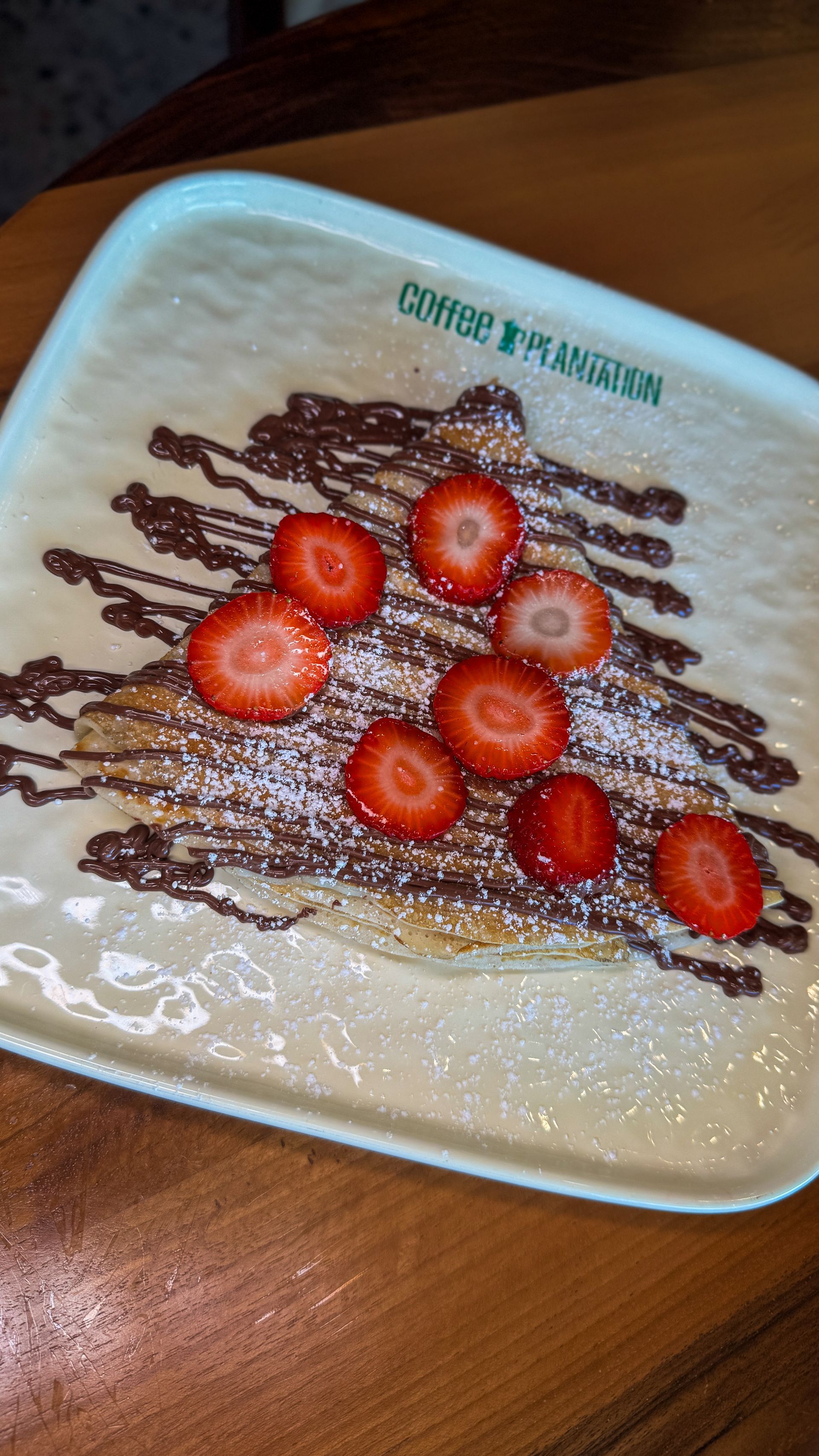 Crepe with chocolate drizzle, strawberry slices, and powdered sugar on a square plate.
