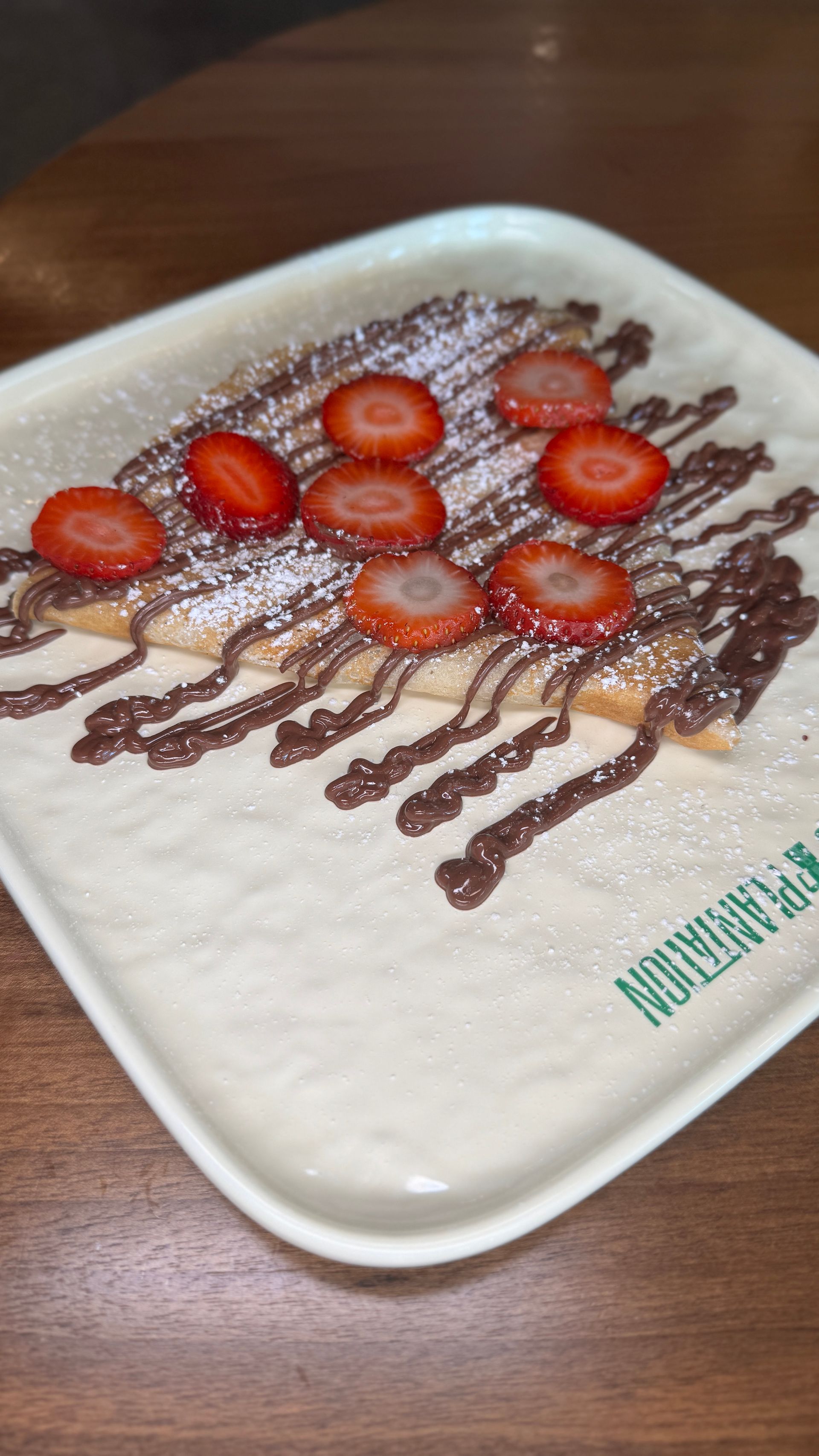 Crepe with chocolate drizzle, strawberries, and powdered sugar on a rectangular plate.