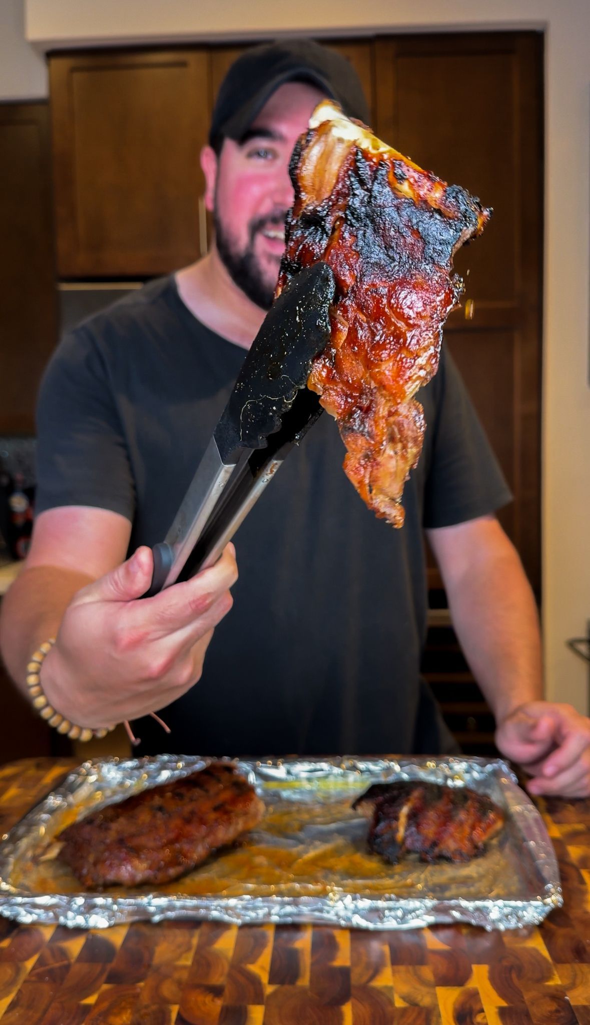 Man holding tongs with a glazed, charred rib over a foil-lined tray of grilled meat indoors