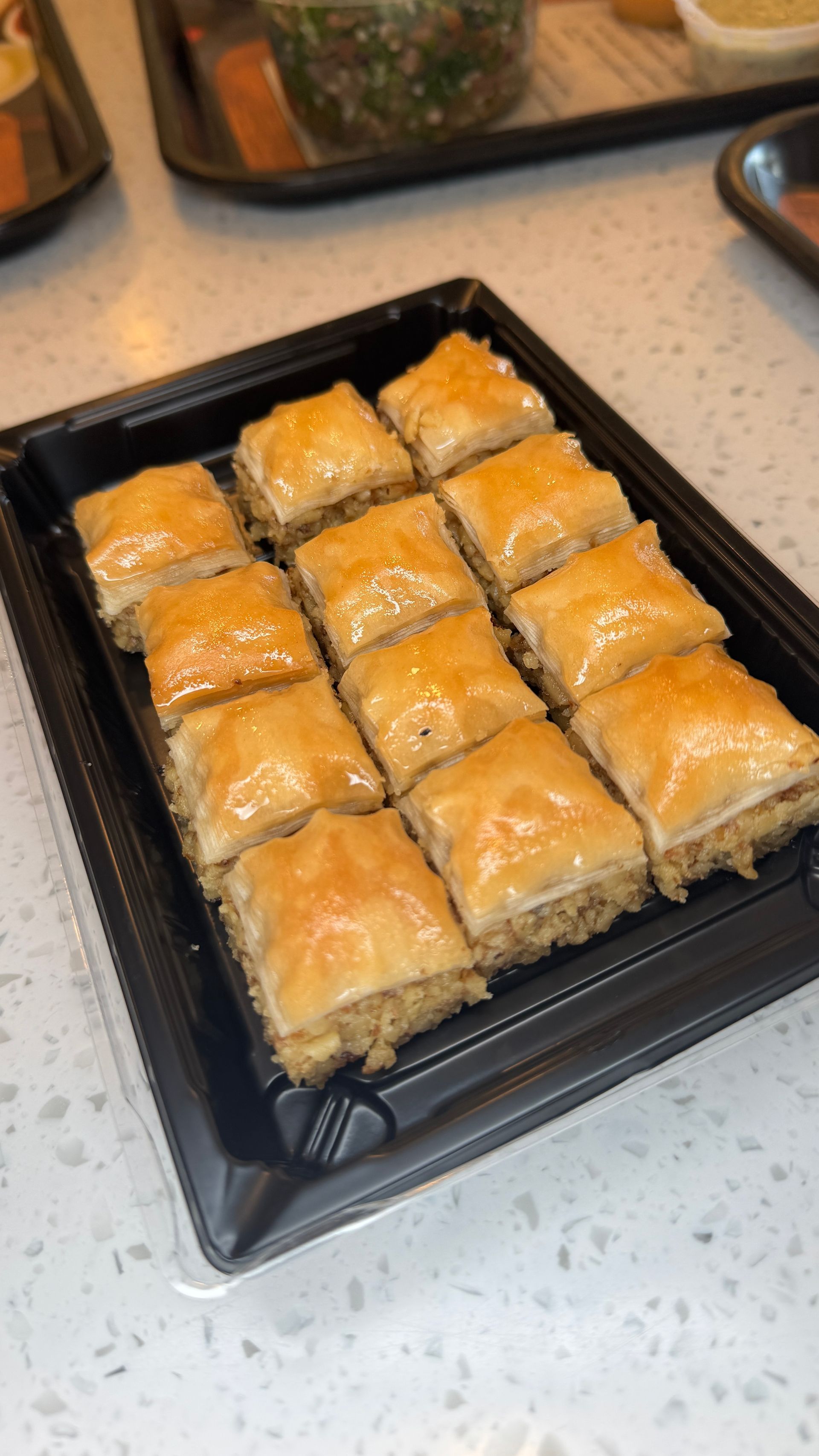 Baklava pastries in a black tray, arranged in rows. The pastries are golden brown and glistening.