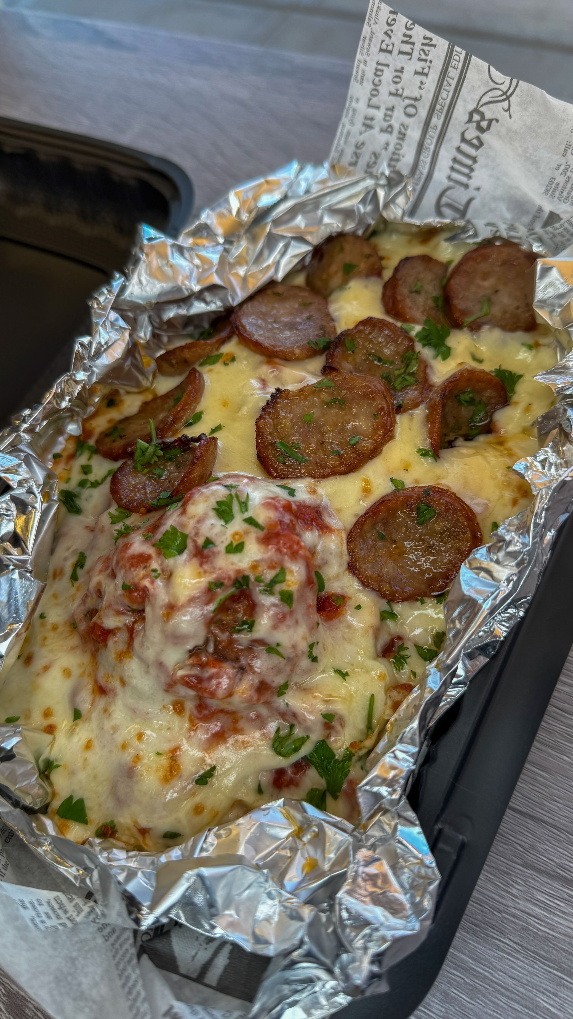 Foil tray of cheesy baked pasta topped with sliced sausage and herbs on a car seat