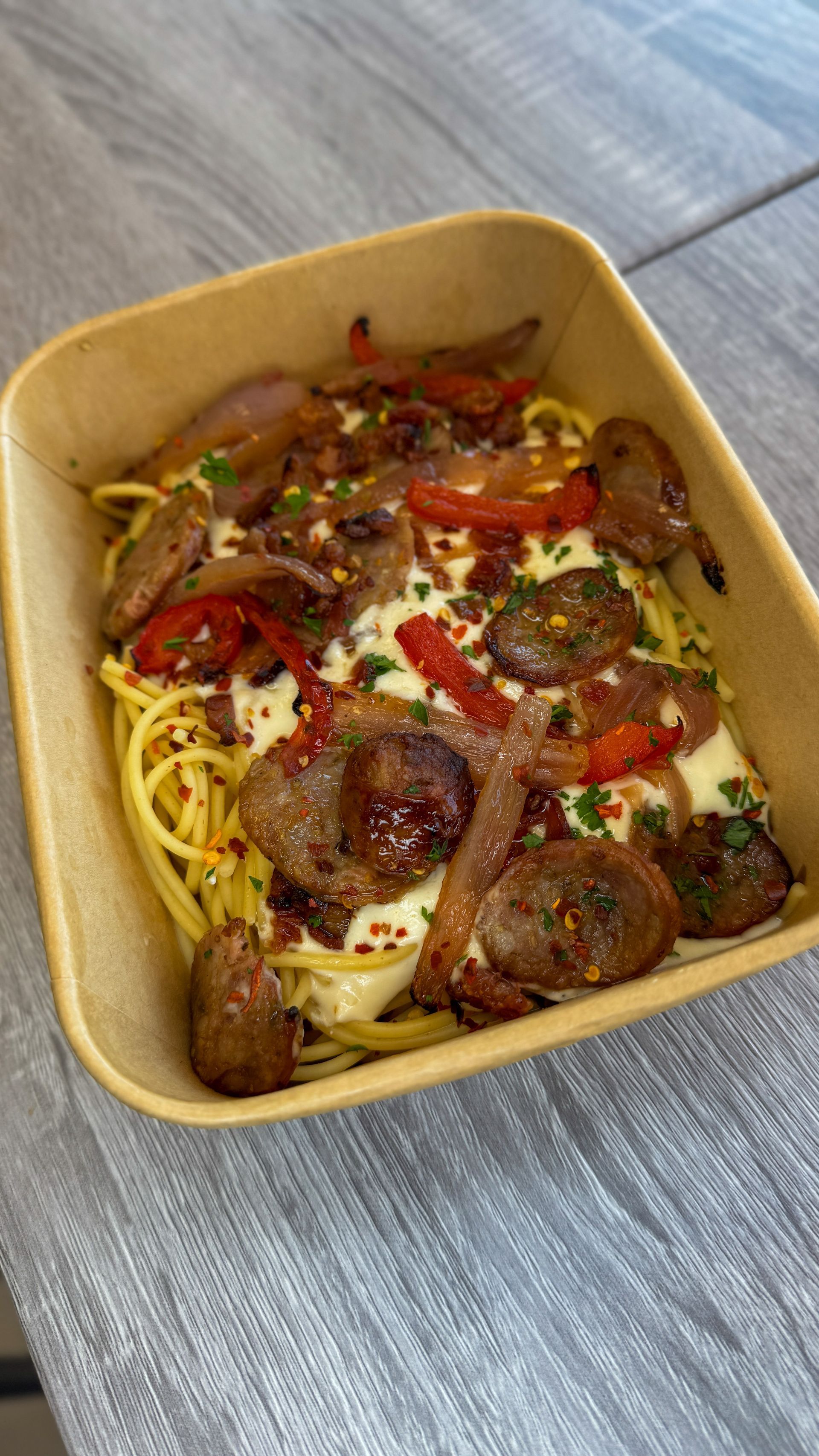 Takeout container of pasta topped with sausage, peppers, herbs, and melted cheese