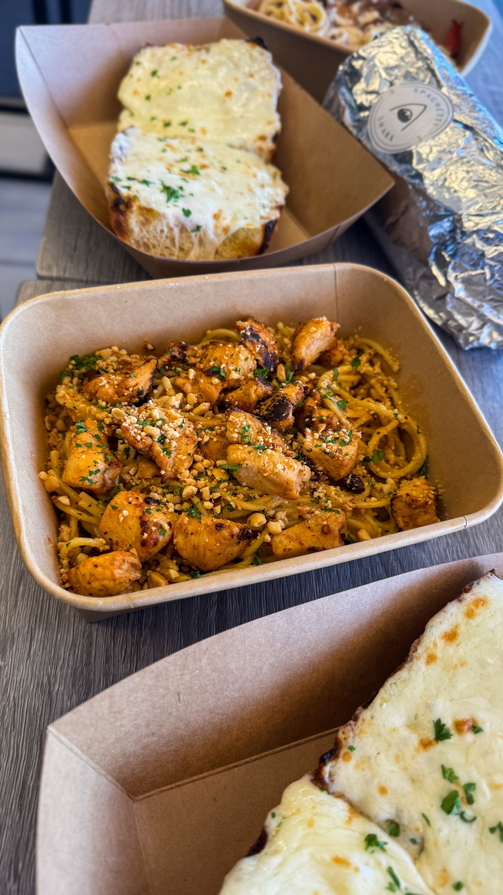 Takeout meal in cardboard containers with pasta, garlic bread, and foil-wrapped breadstick