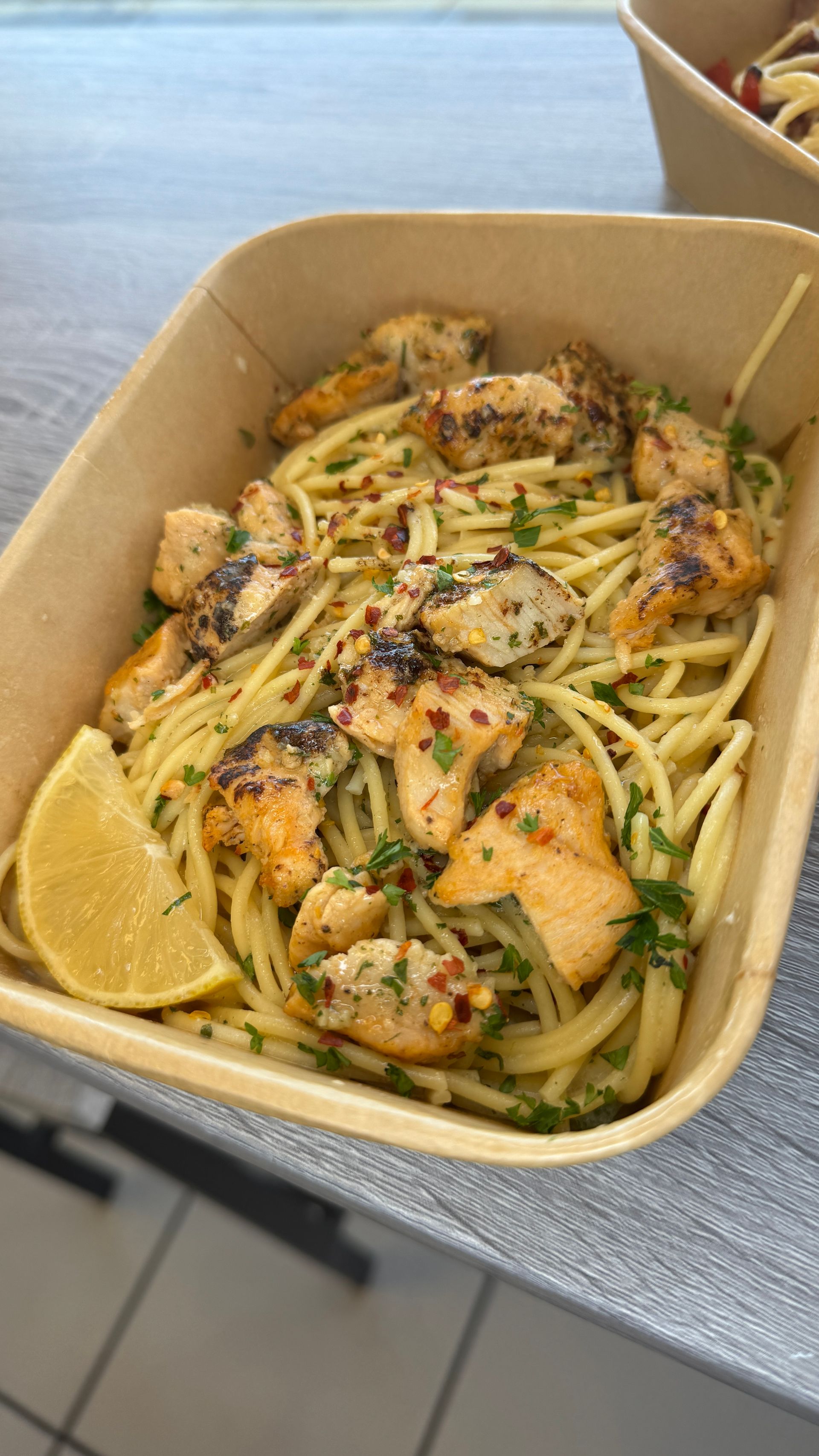 Takeout pasta with grilled chicken, lemon wedges, and herbs in a cardboard container