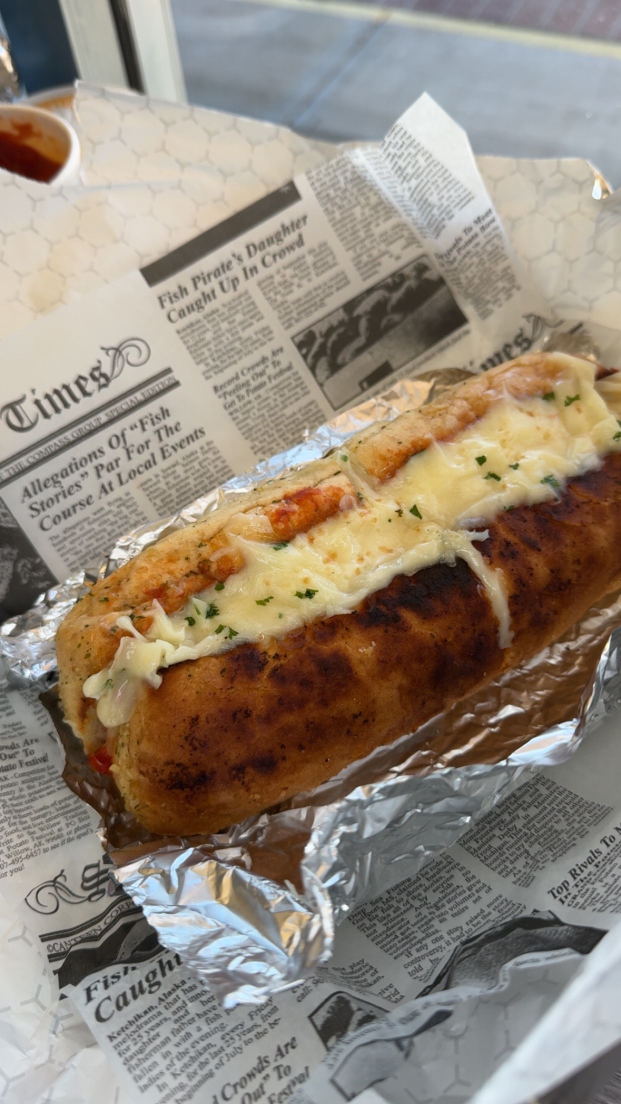 Hot dog in a bun topped with melted cheese and herbs, wrapped in foil on newspaper-lined tray.
