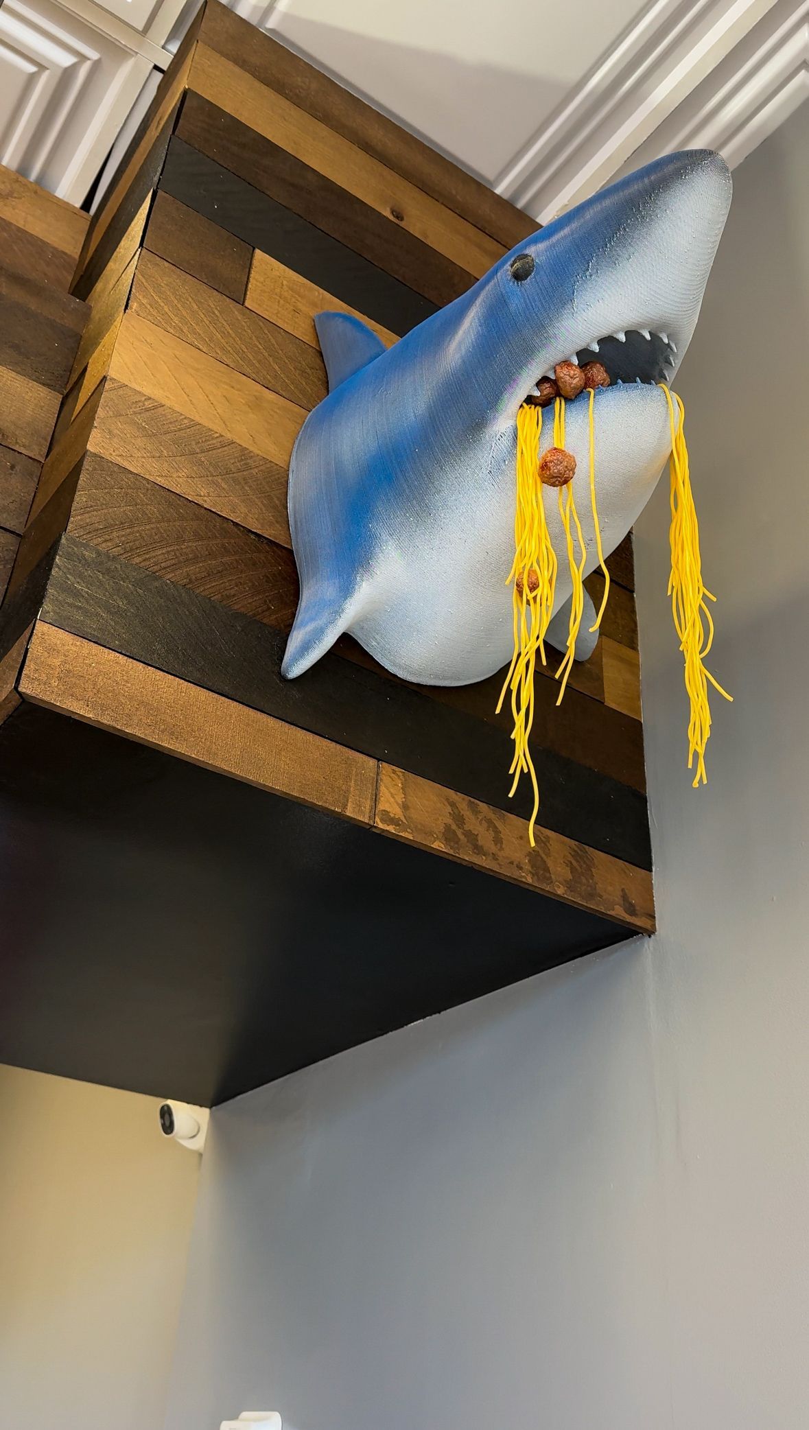 Blue shark decoration hanging from a wooden shelf with yellow streamers against a gray wall