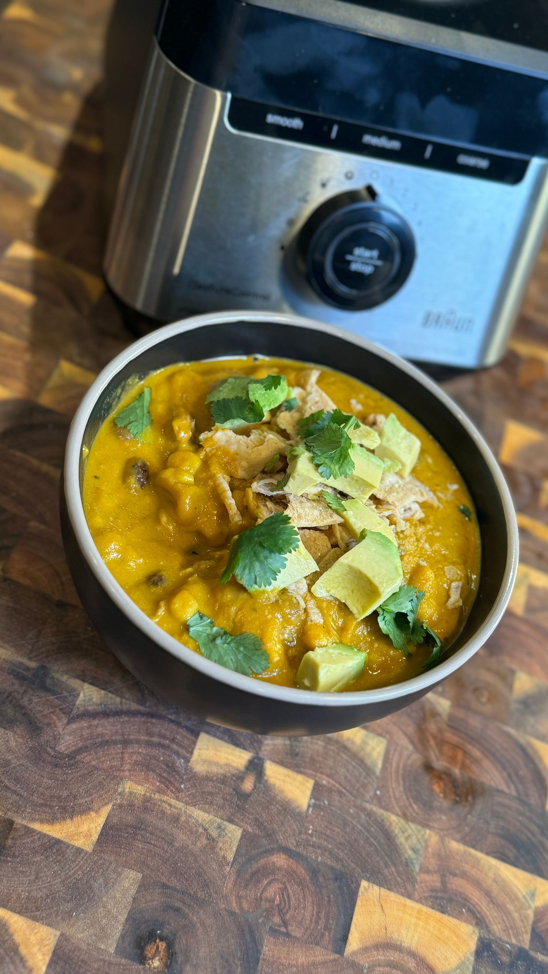 A bowl of soup with avocado and cilantro next to a blender on a wooden table.