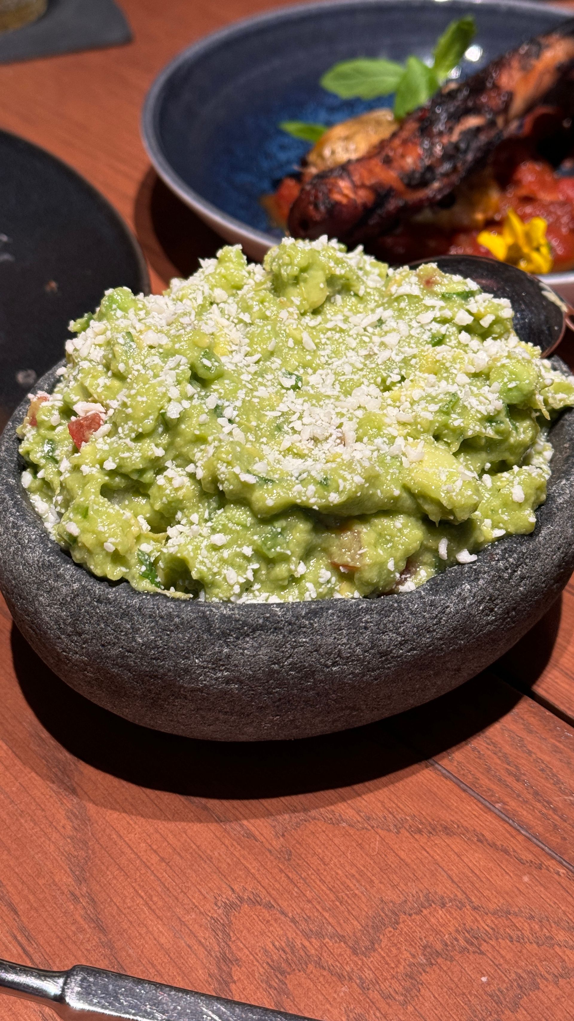 A bowl of guacamole is sitting on a table next to a plate of food.