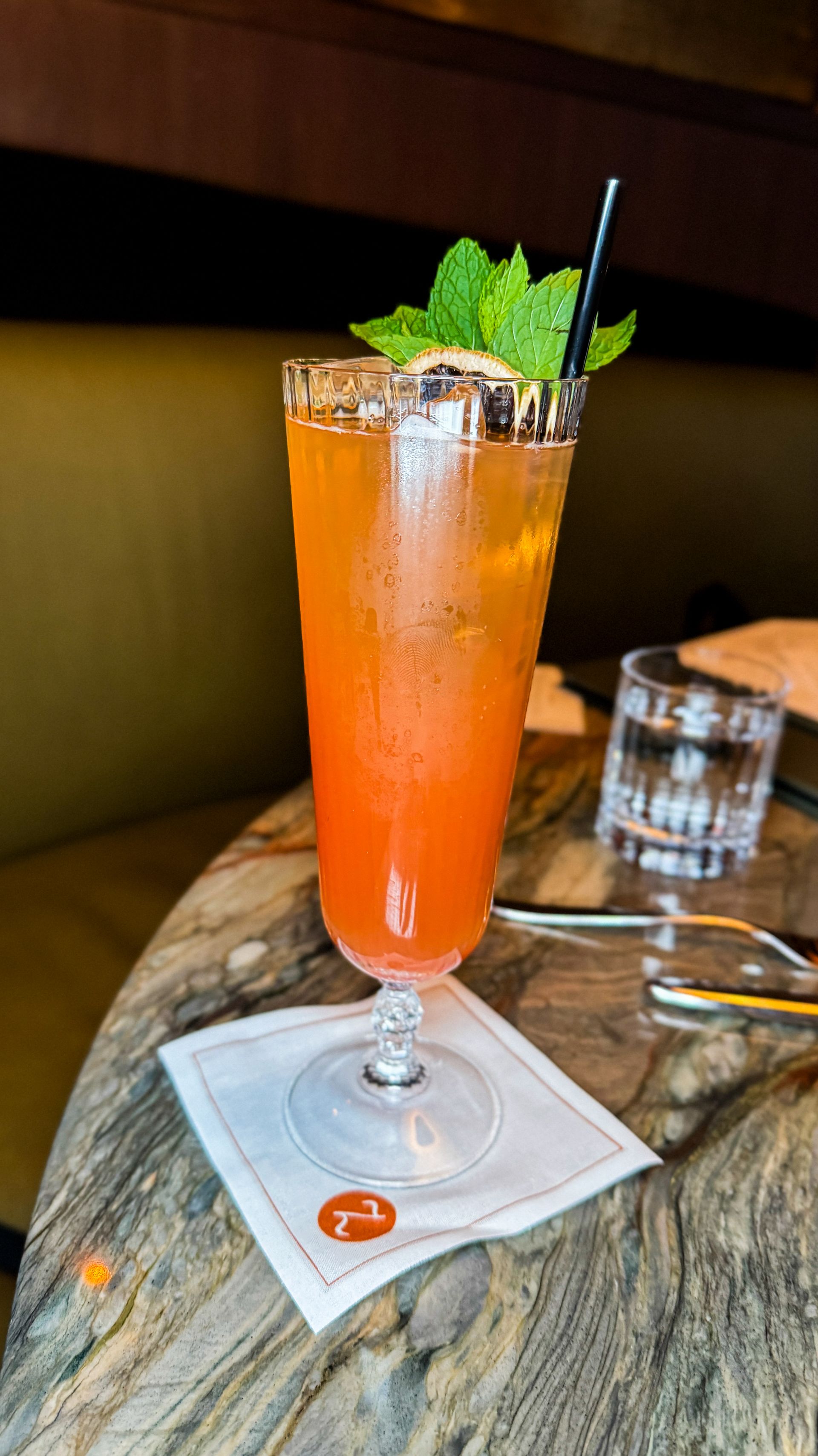 Tall orange cocktail with mint and a straw on a marble table in a dim bar.