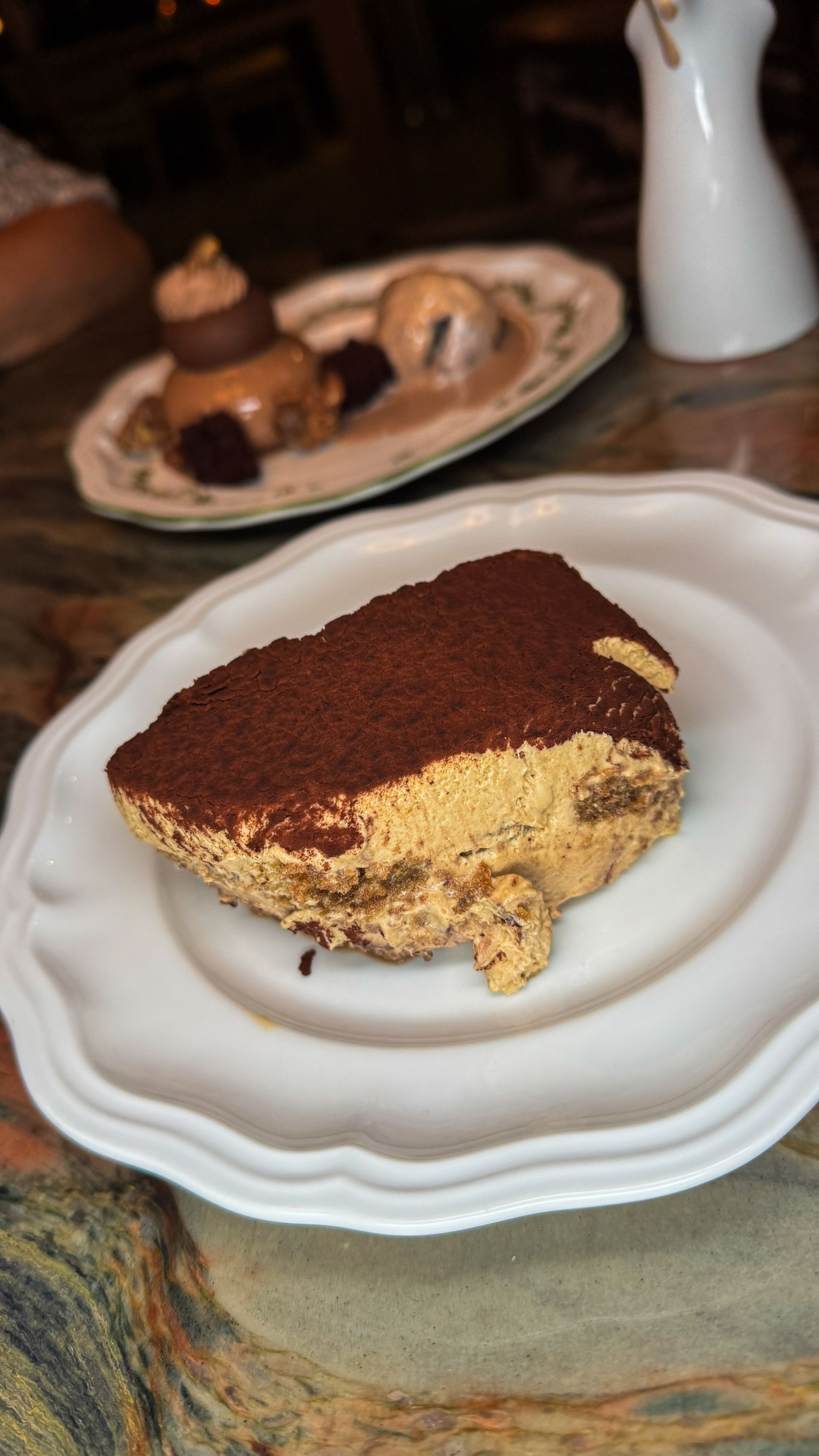 Slice of tiramisu on a white plate with a cup of coffee in the background