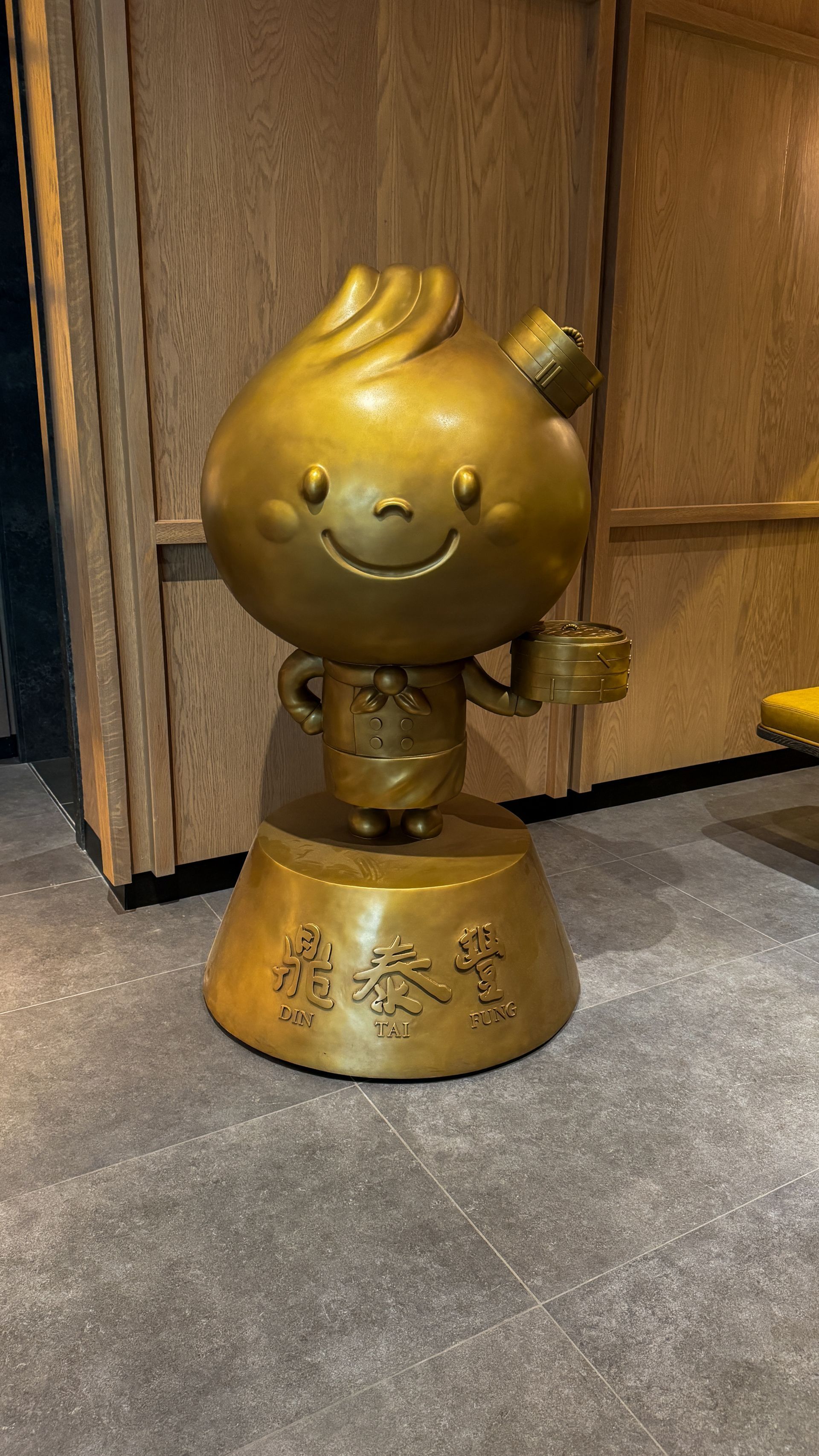 Gold-colored cartoon figurine on a base, smiling with hands on hips, displayed on a floor by wooden cabinets