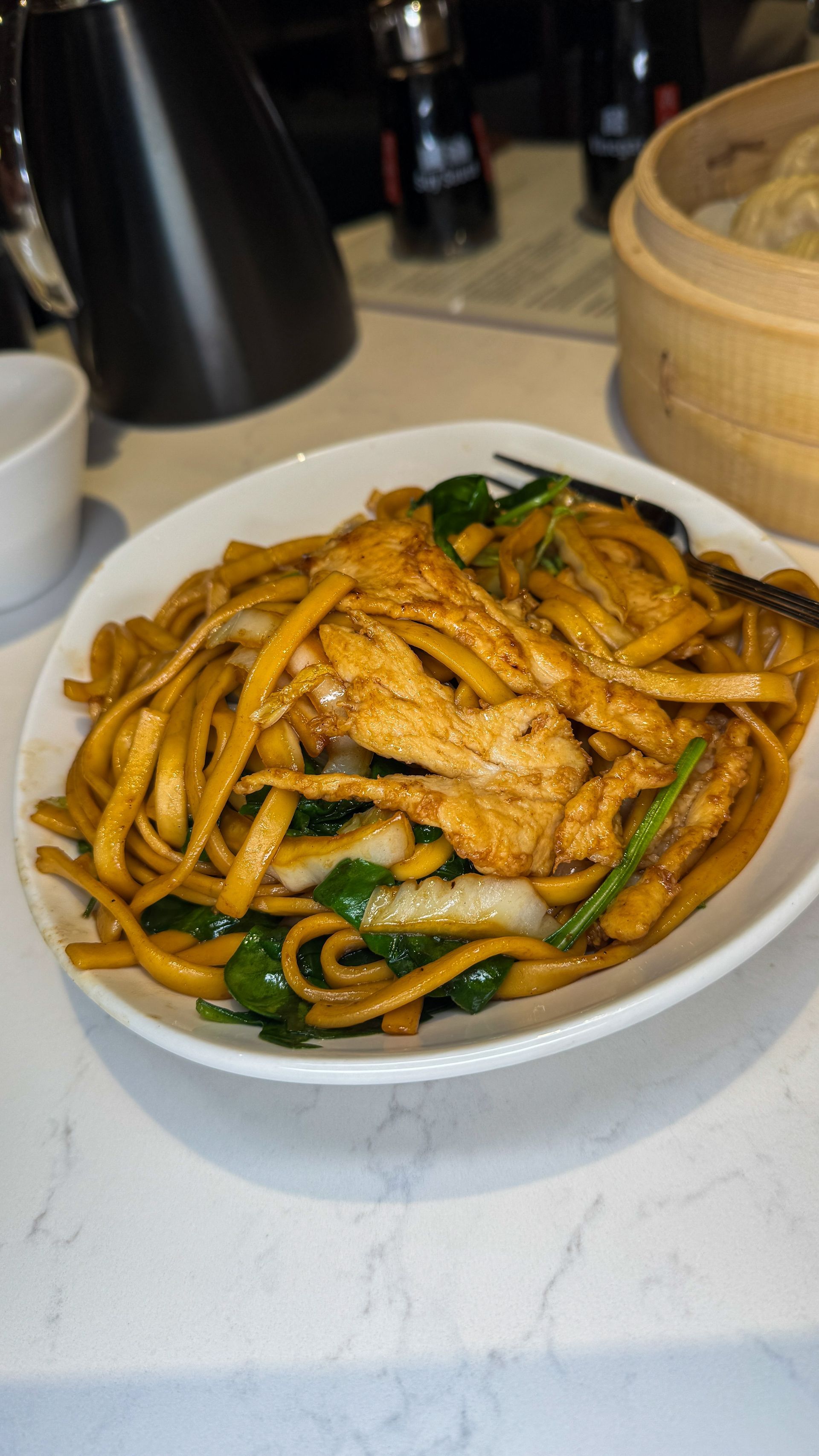Bowl of stir-fried noodles with vegetables and meat on a table, with a bamboo steamer nearby