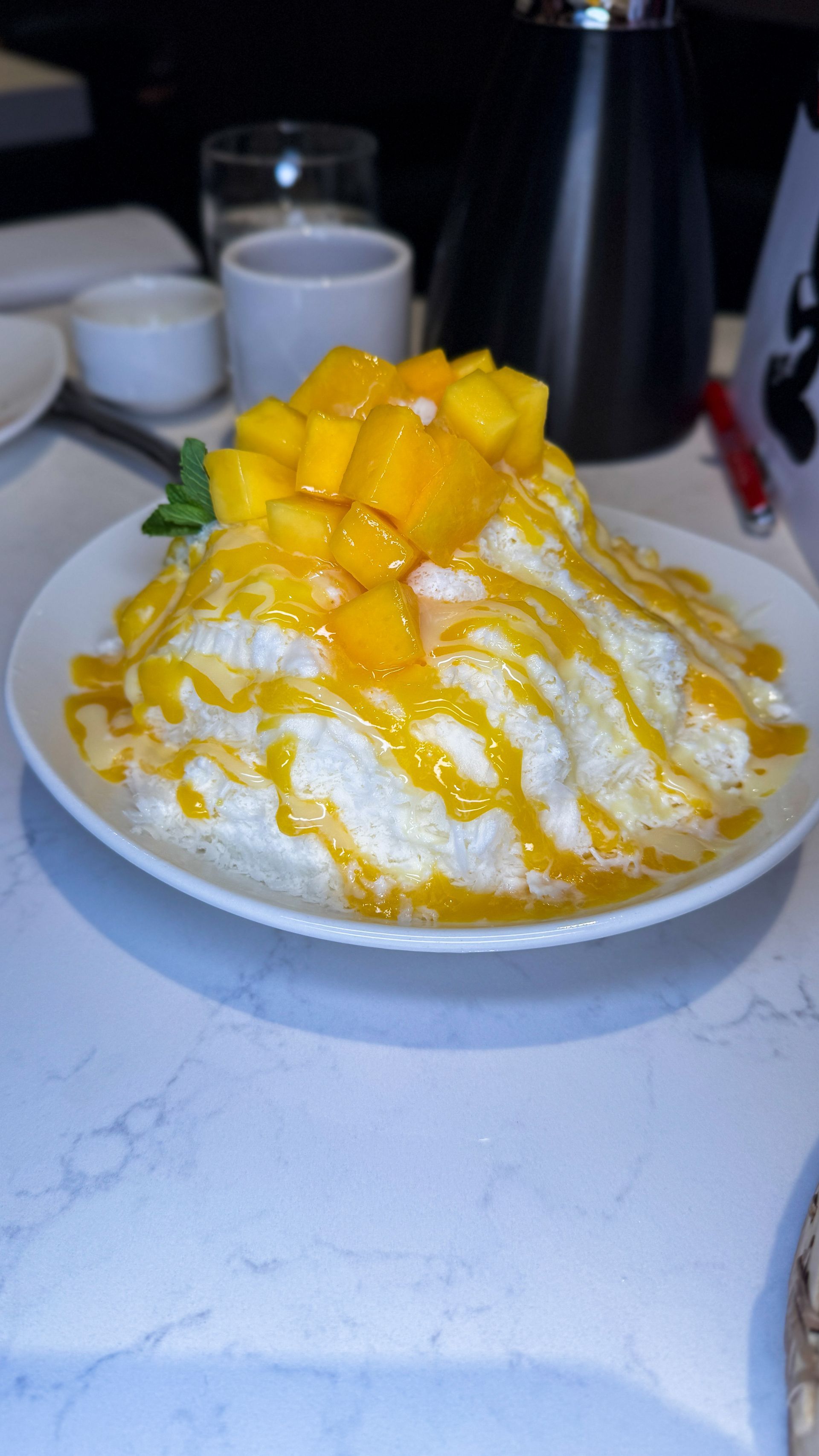 Mango shaved ice dessert topped with syrup on a white plate in a café setting