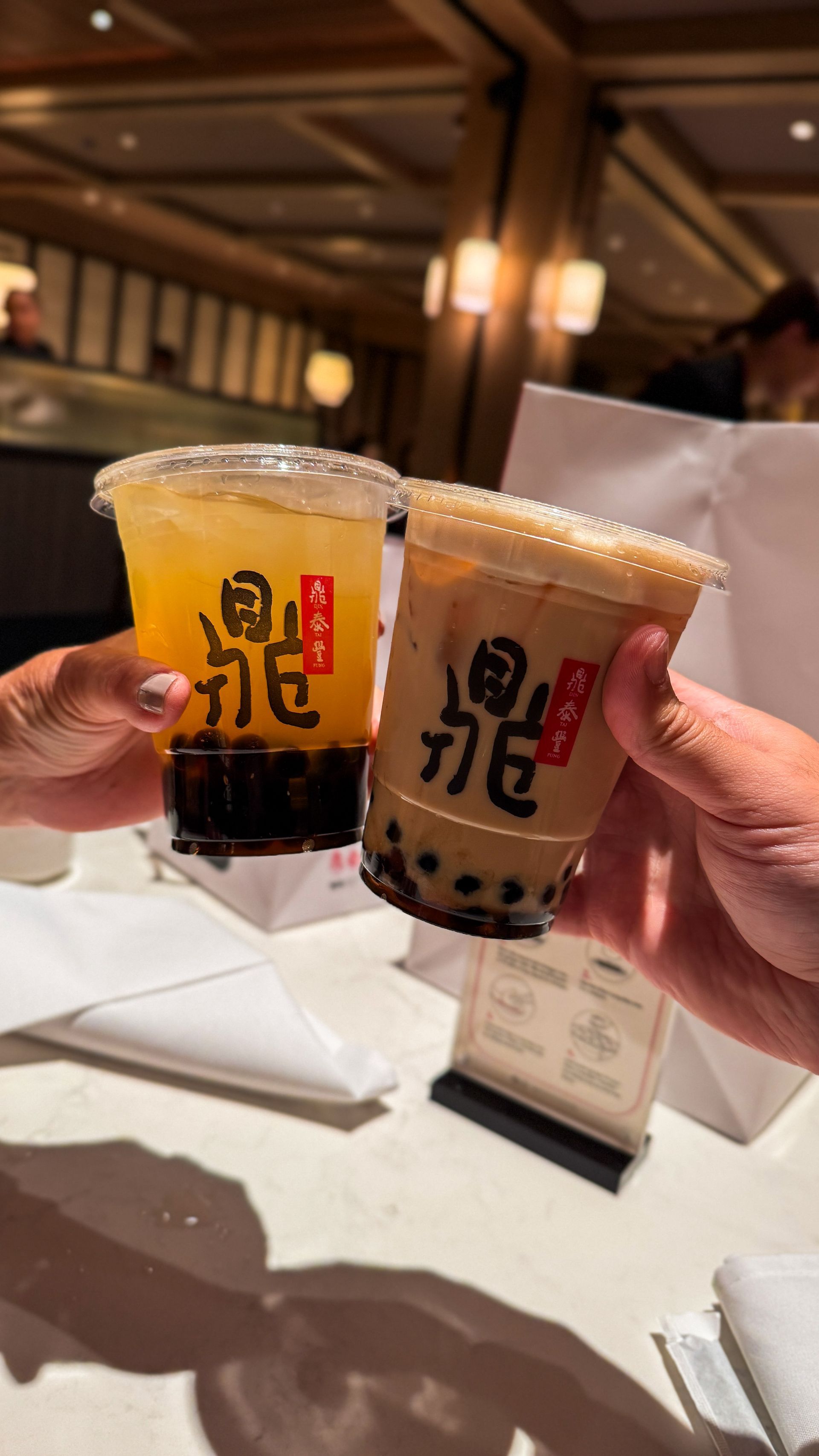 Two hands holding two iced drinks with black and red labels in a warmly lit restaurant.