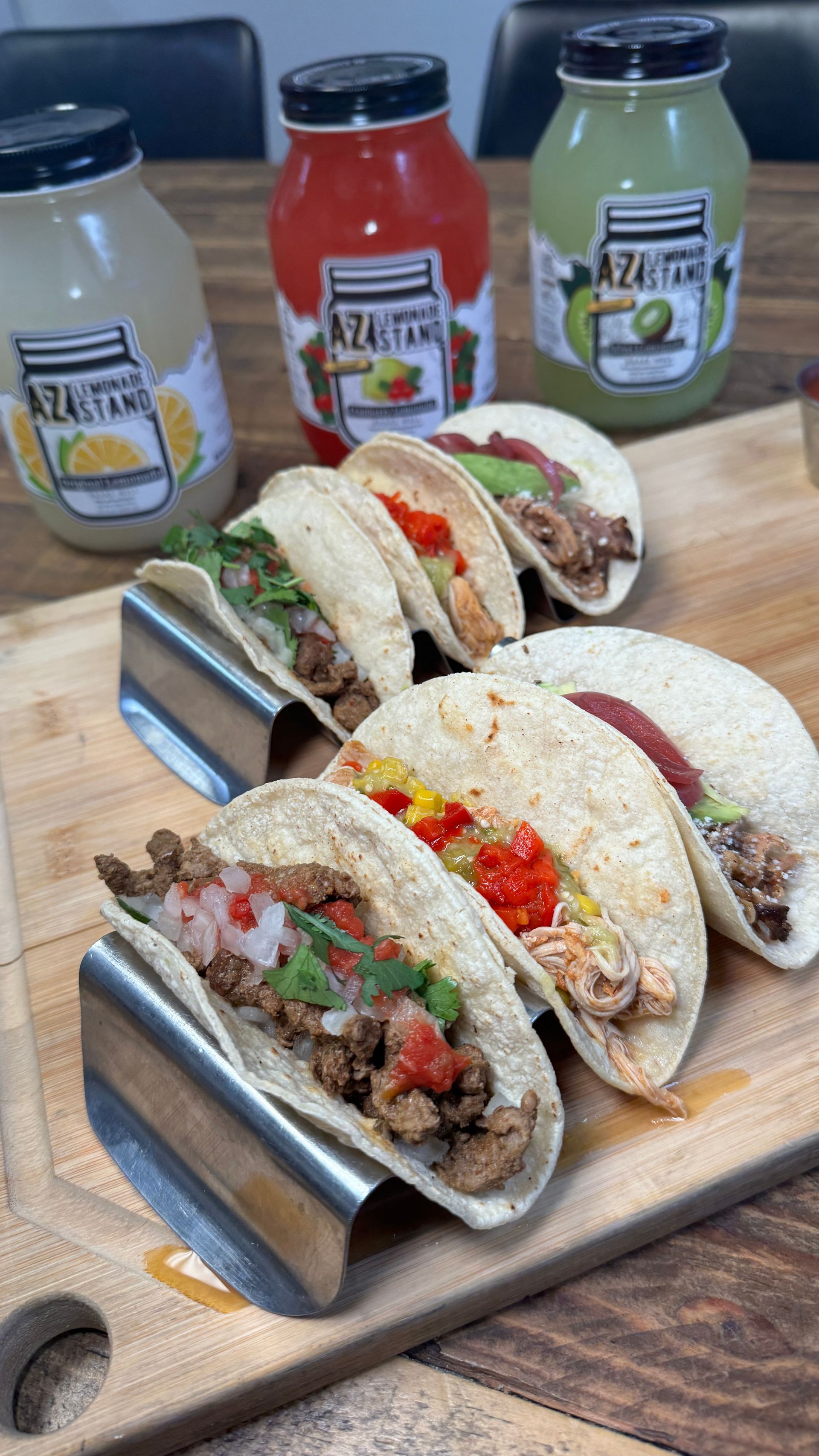 Three tacos are sitting on a wooden cutting board next to bottles of sauce.