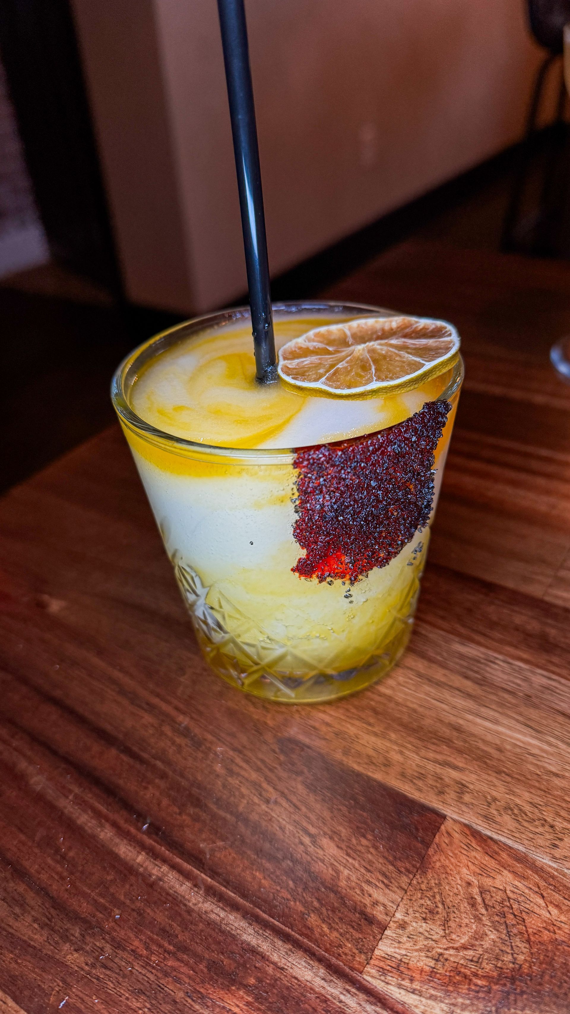 Yellow cocktail with orange slice and blackberry garnish in a short glass on a wooden table