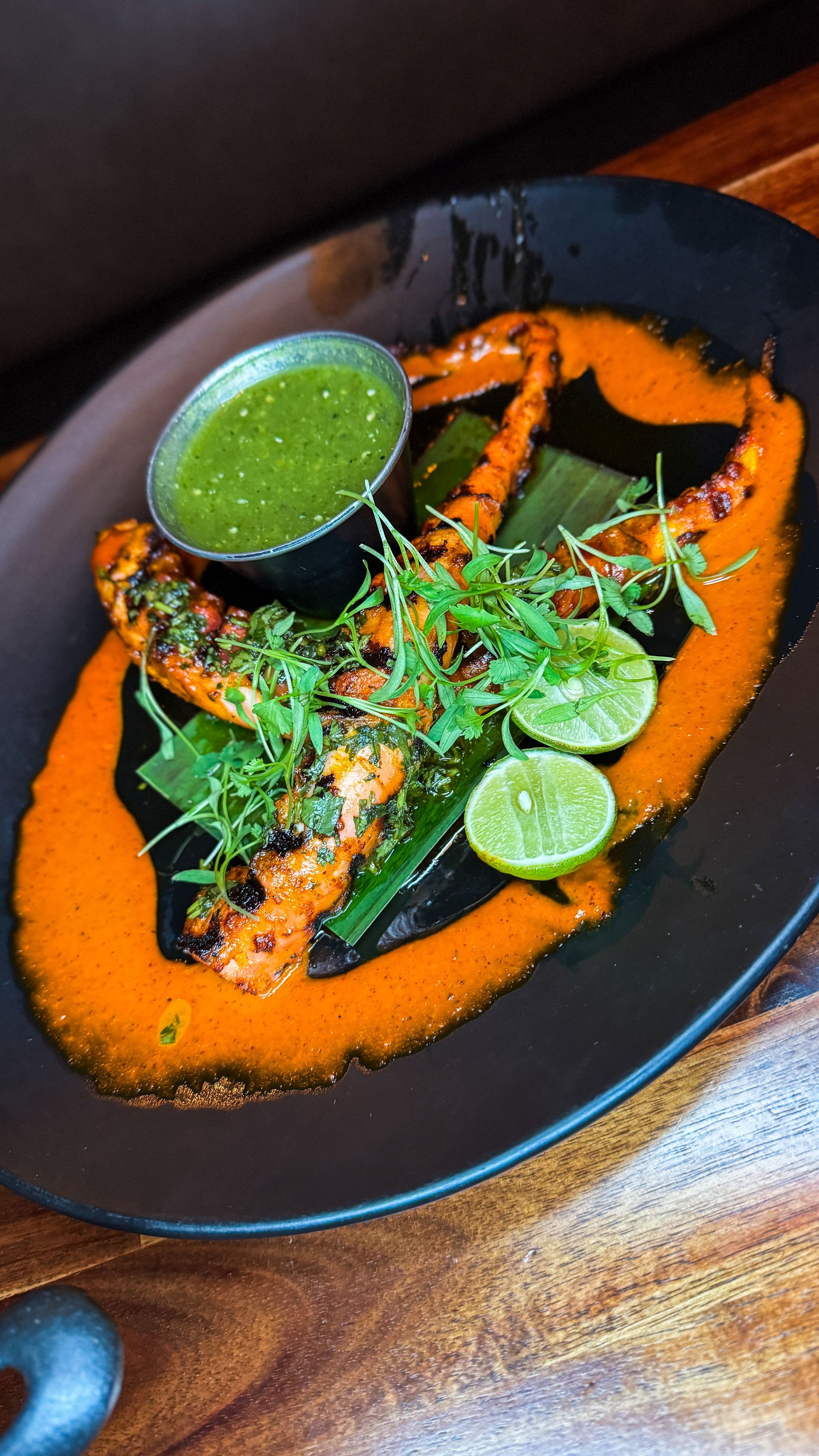 Plated orange curry with green chutney, herbs, and lime wedges on a black dish
