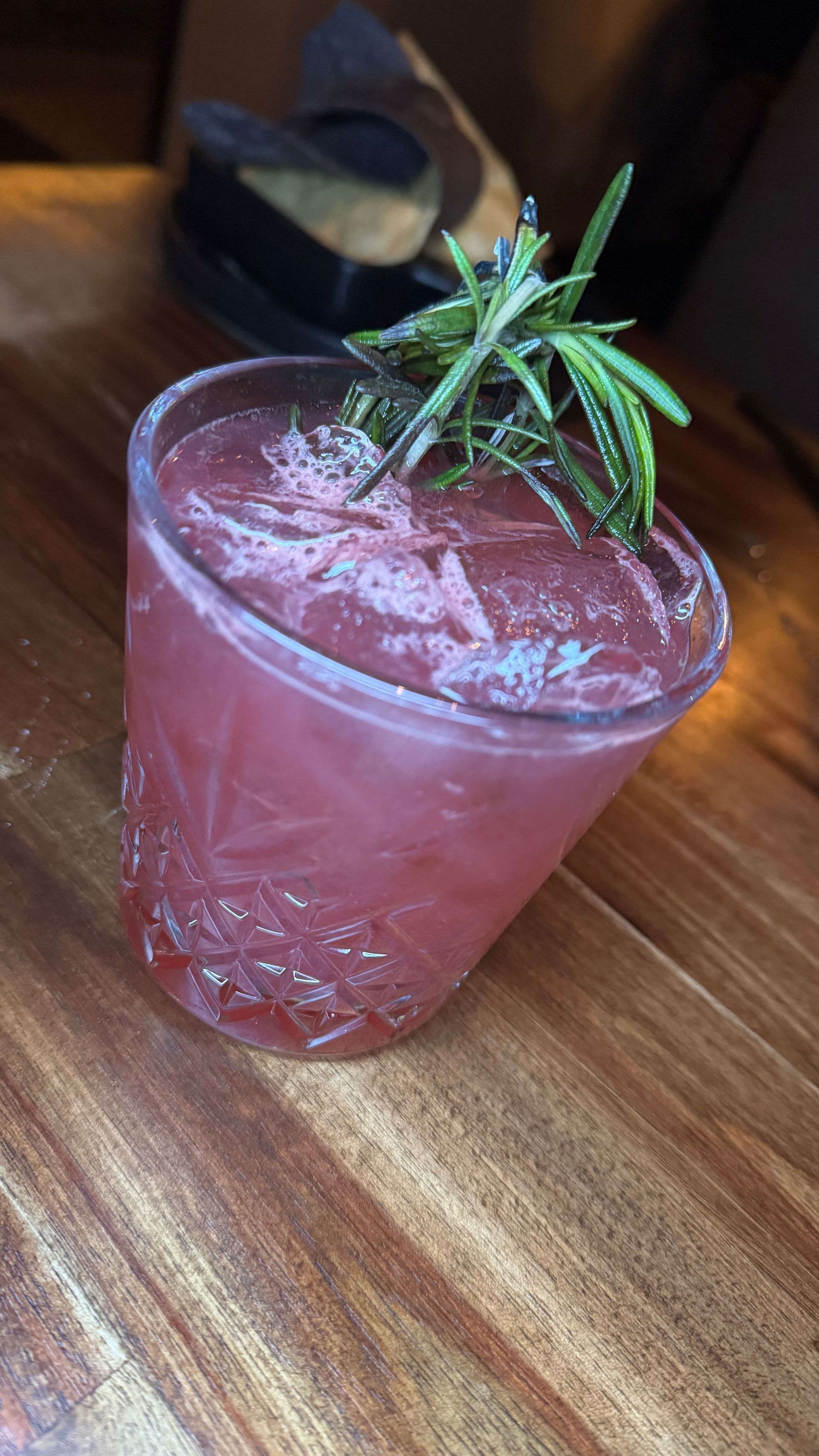 Pink cocktail in a glass with ice and rosemary garnish on a wooden table