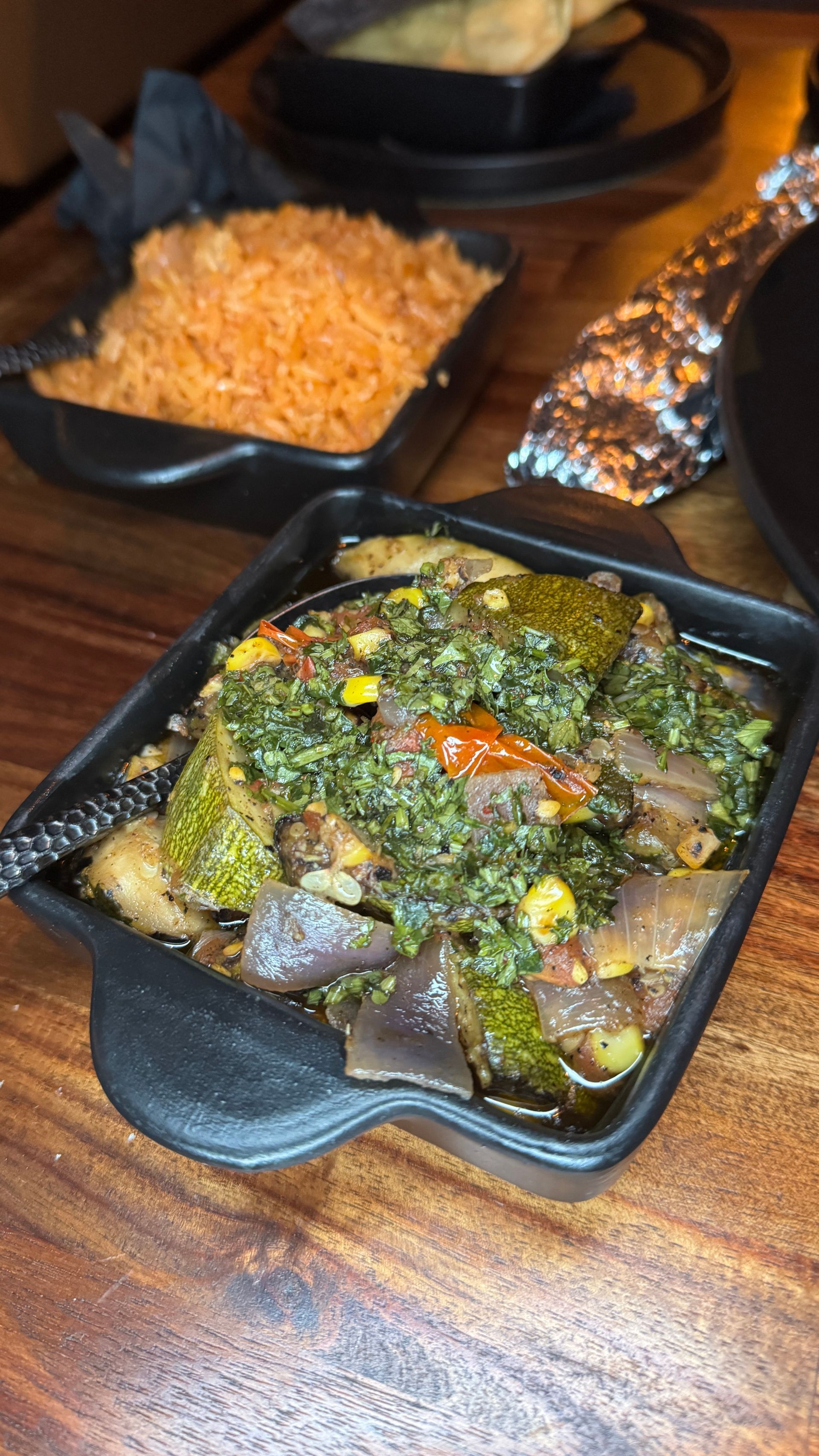 Takeout food on a table: black container with green vegetable dish, orange rice, and foil-wrapped item