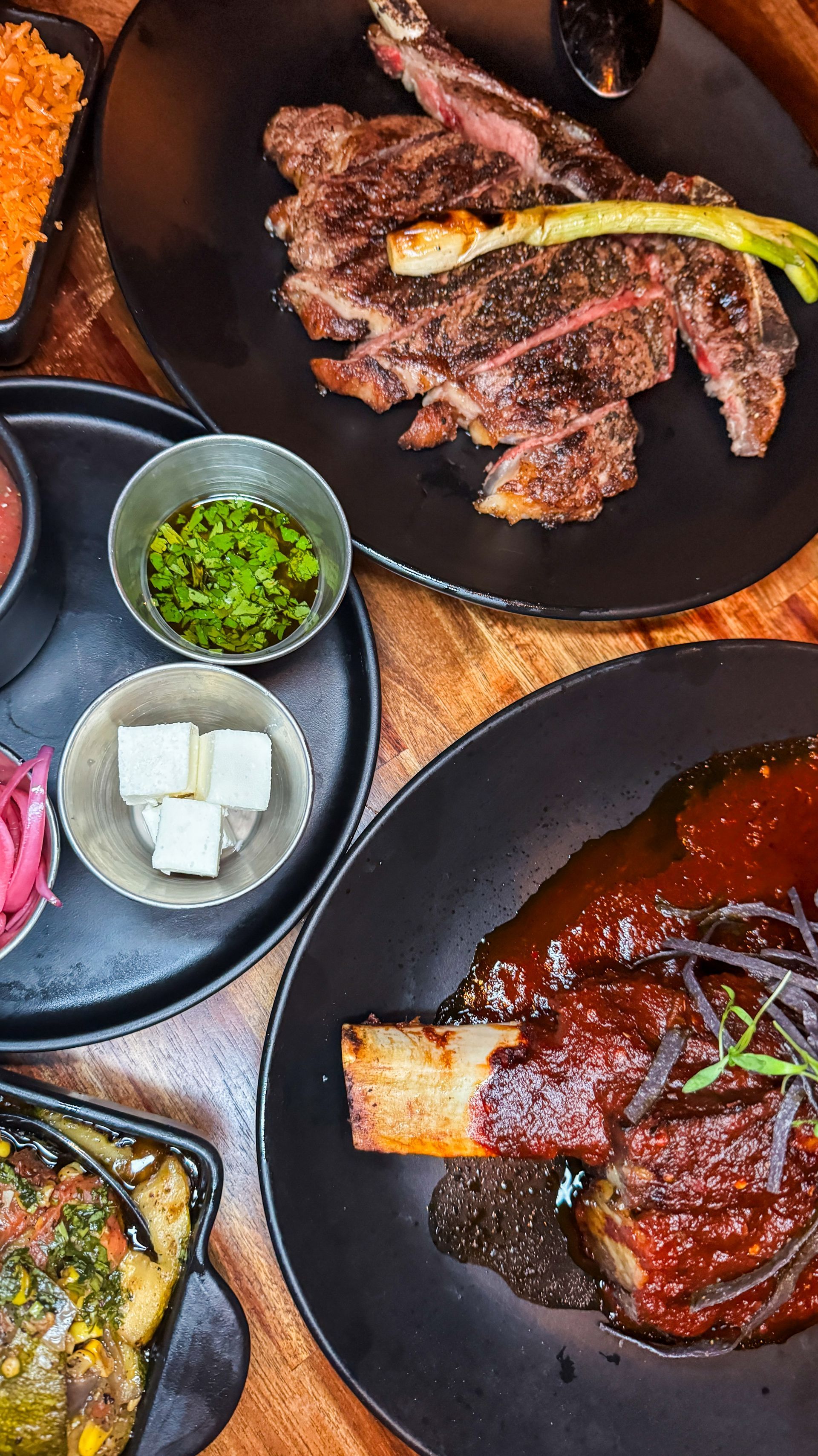 Assorted grilled meats and side dishes on black plates, including sauce, onions, and cubes of tofu.
