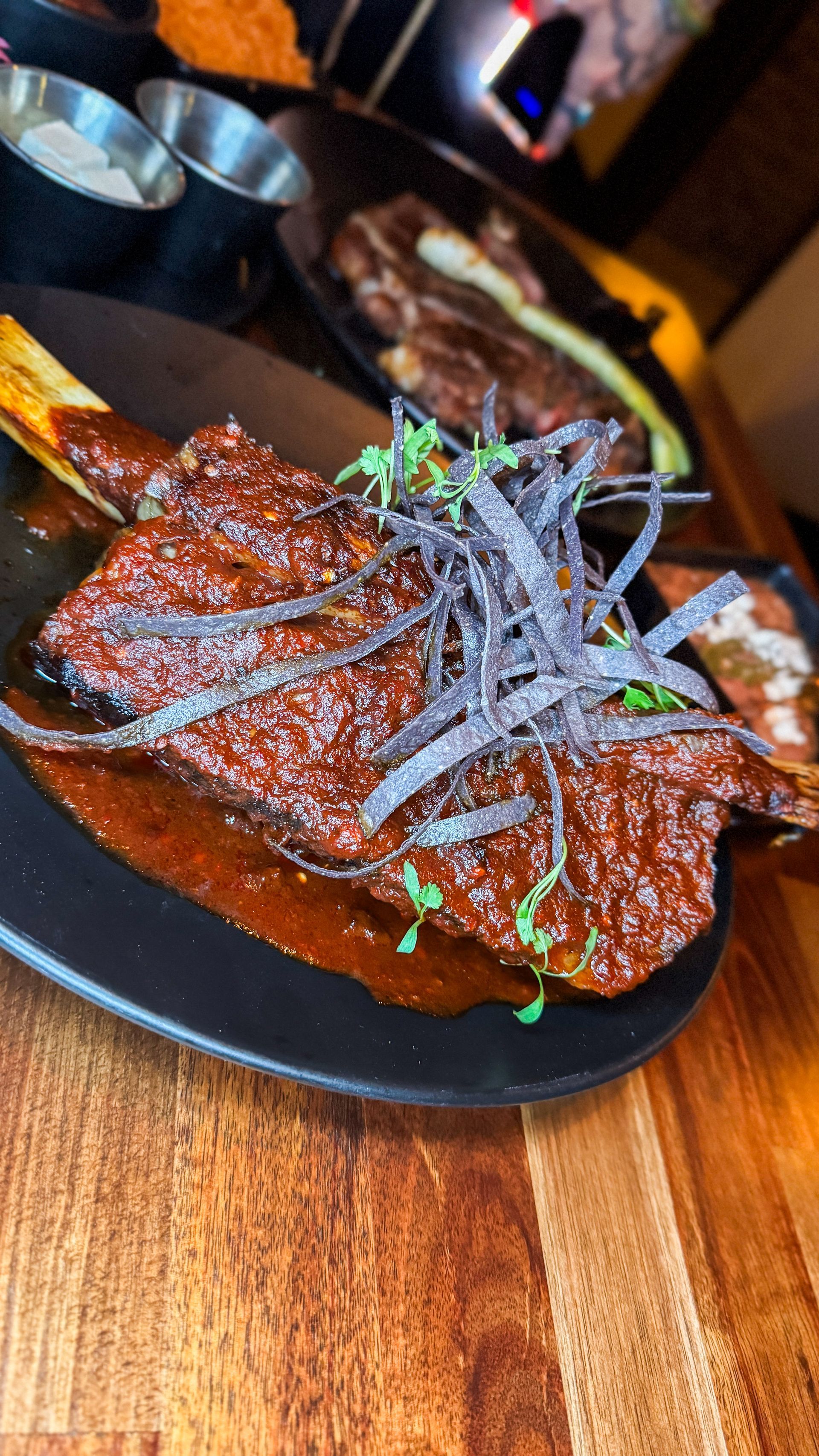Glazed grilled meat on a black plate, topped with herbs, on a wooden table.