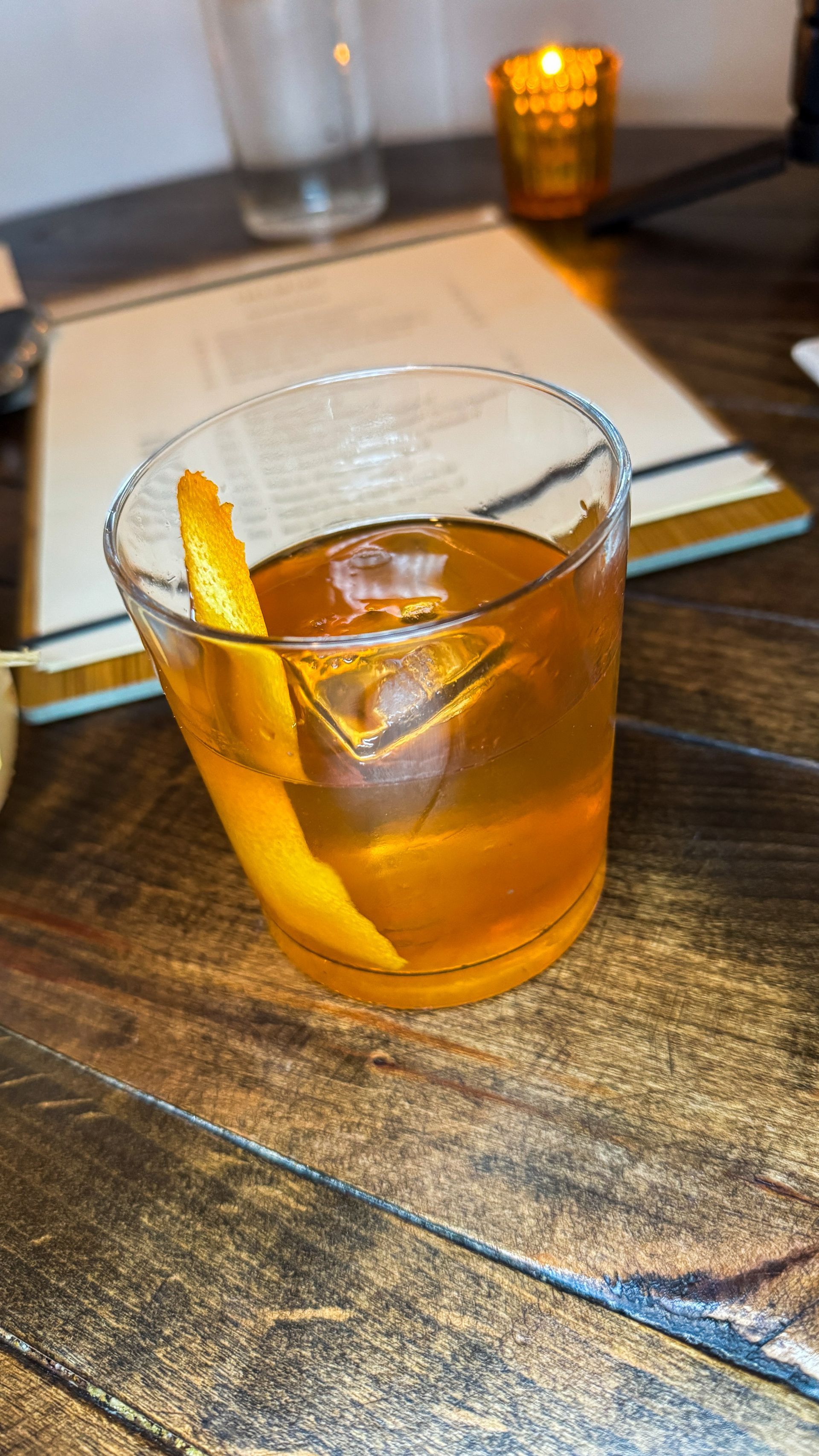 Amber cocktail with orange peel in a low glass on a wooden table, lit by warm candlelight.
