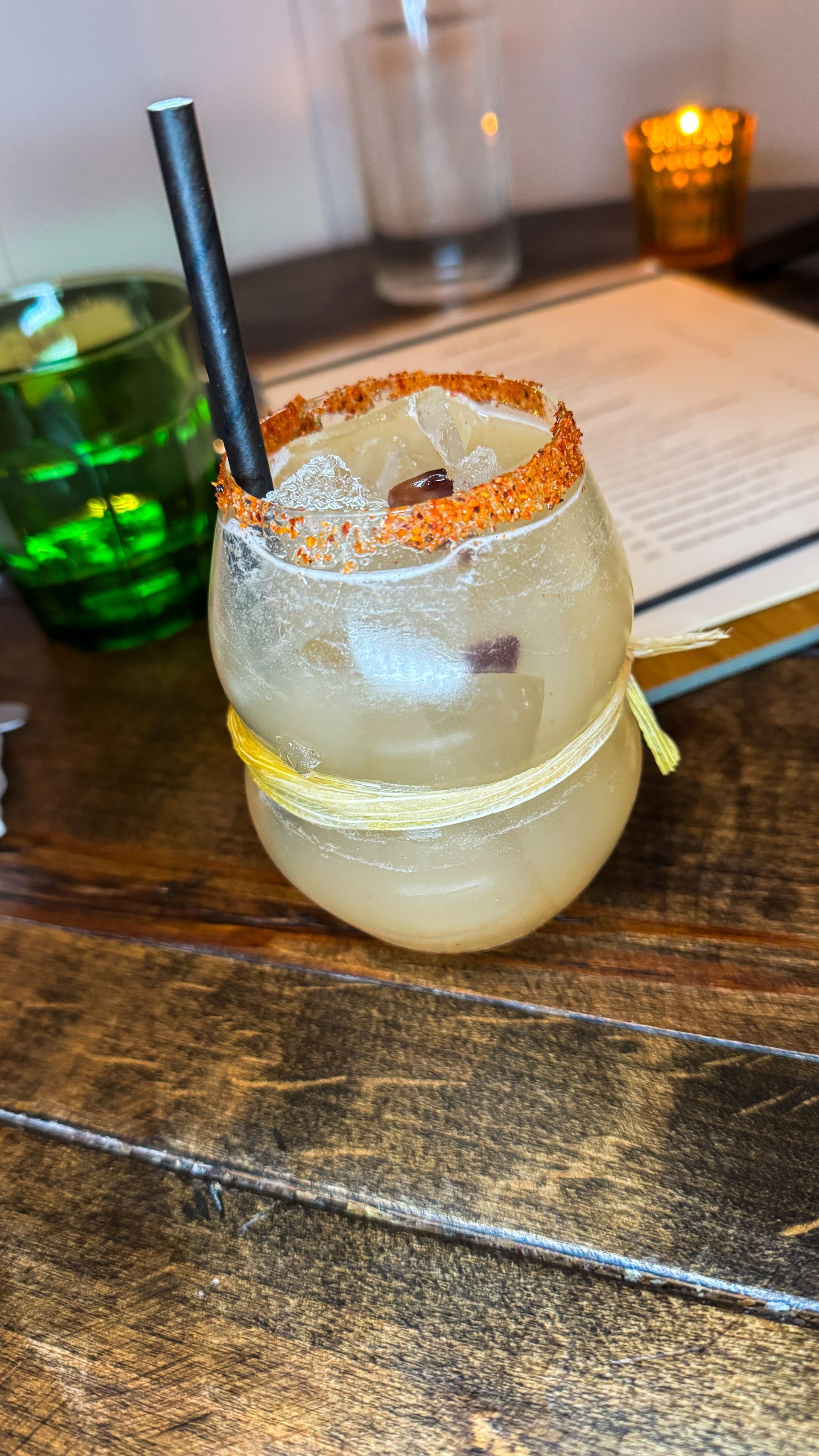 Salt-rimmed cocktail with a black straw on a wooden bar, with green bottle and candlelight in the background