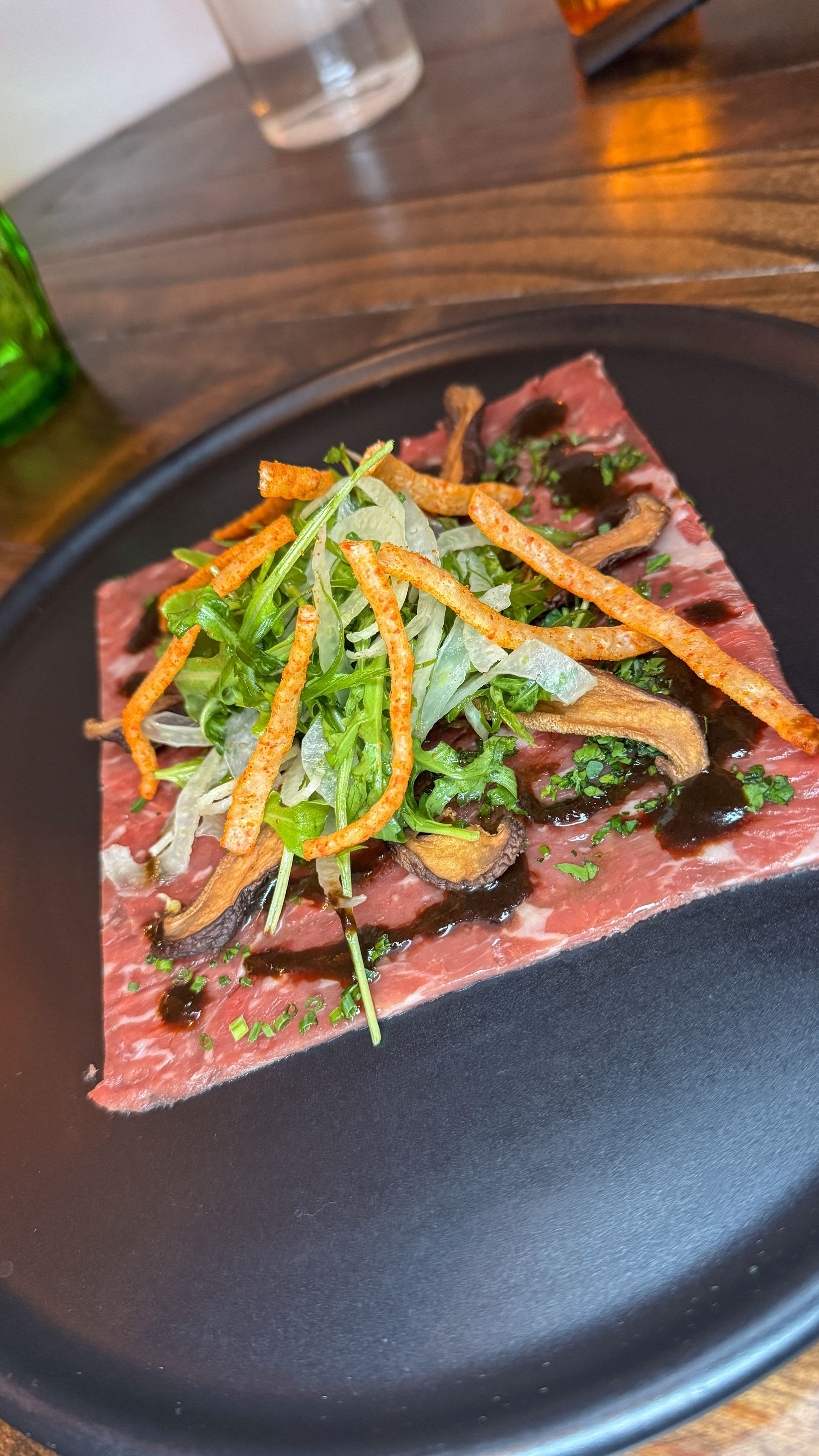 A plated square tart with greens and shredded vegetables on a black plate at a wooden table