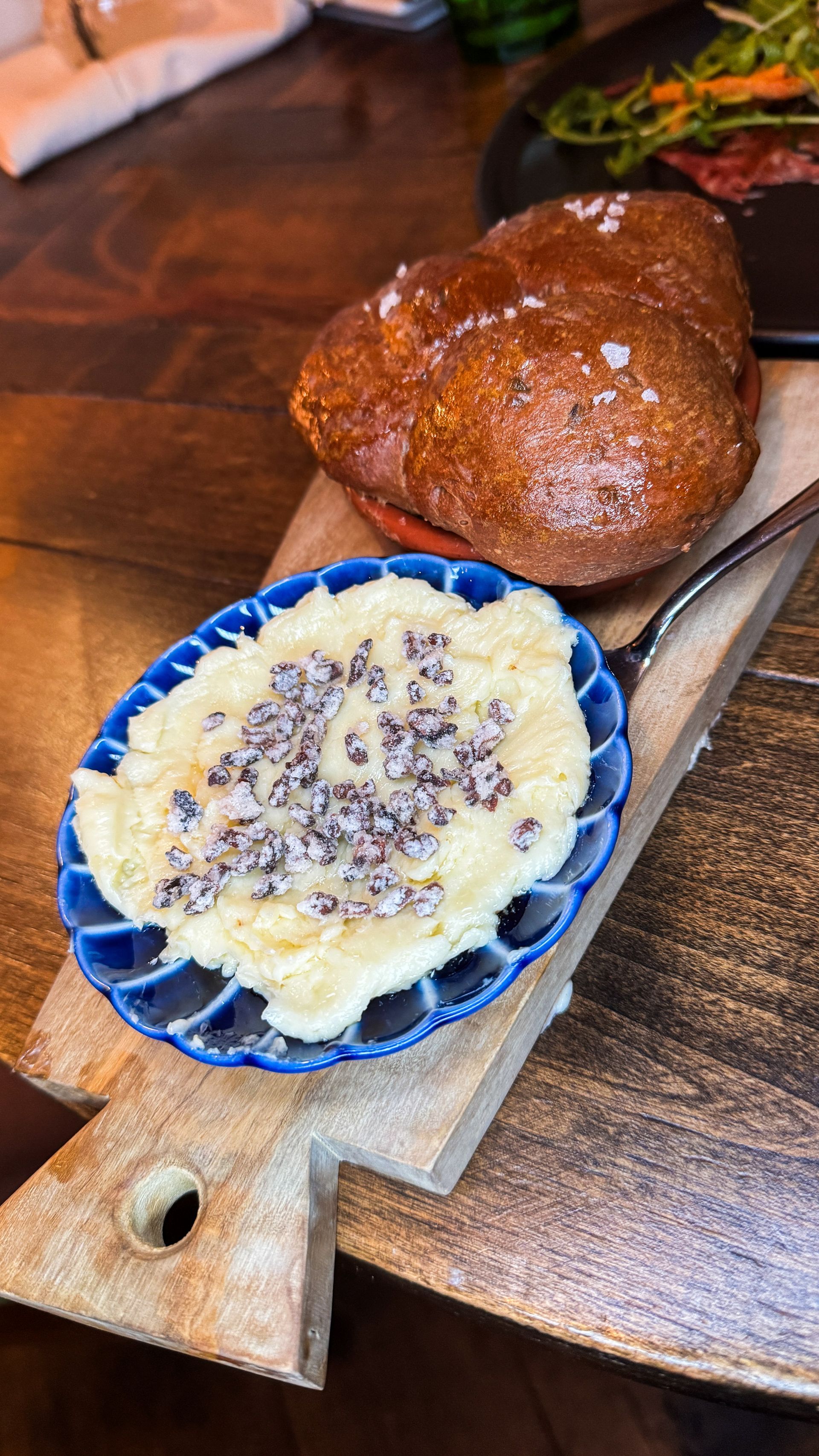 Blue dish of creamy dessert with sprinkled topping on a wooden serving board beside a glazed bread roll