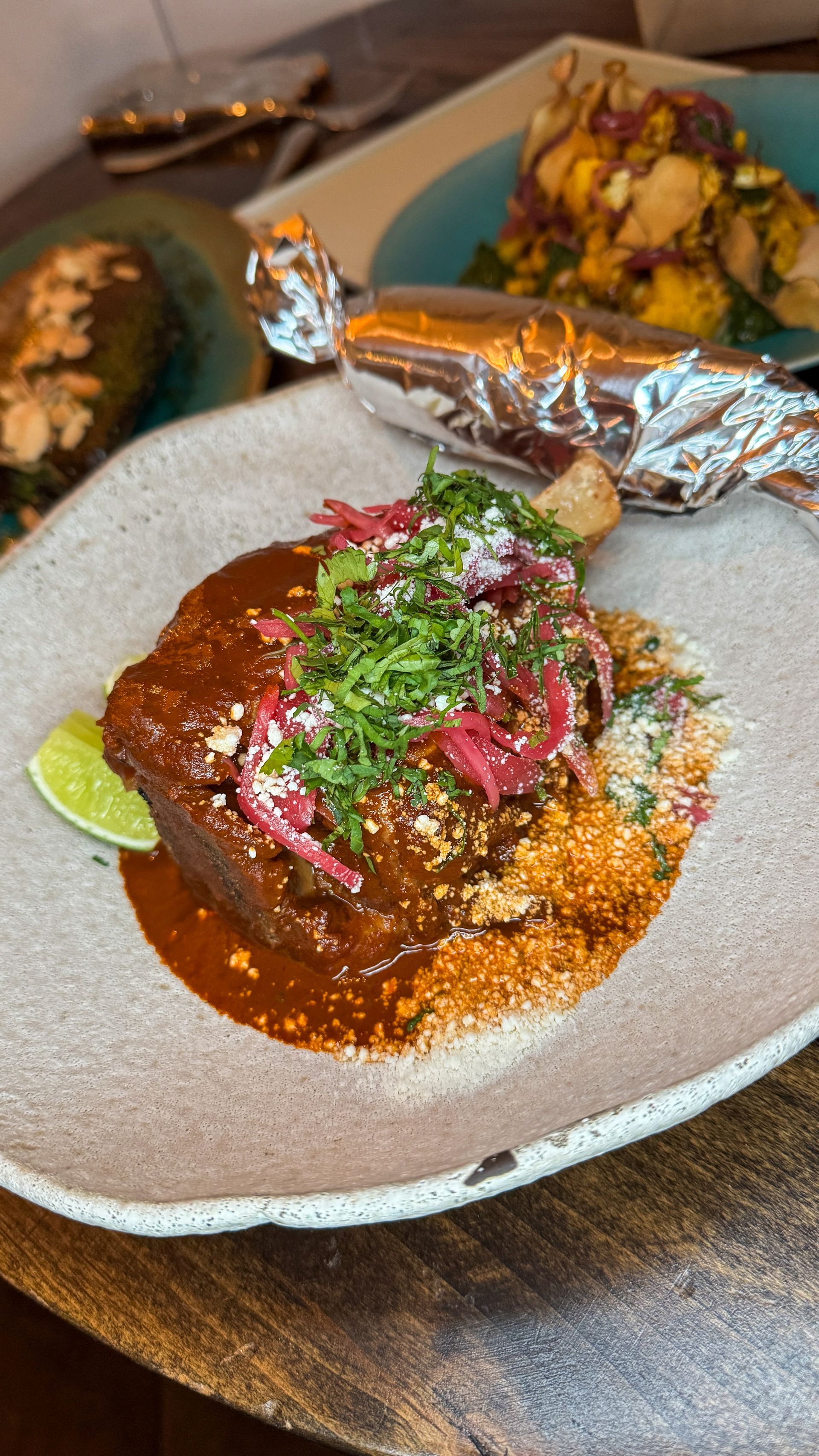 Plated saucy meat dish garnished with herbs and pink onions, with foil-wrapped sides on a dark table