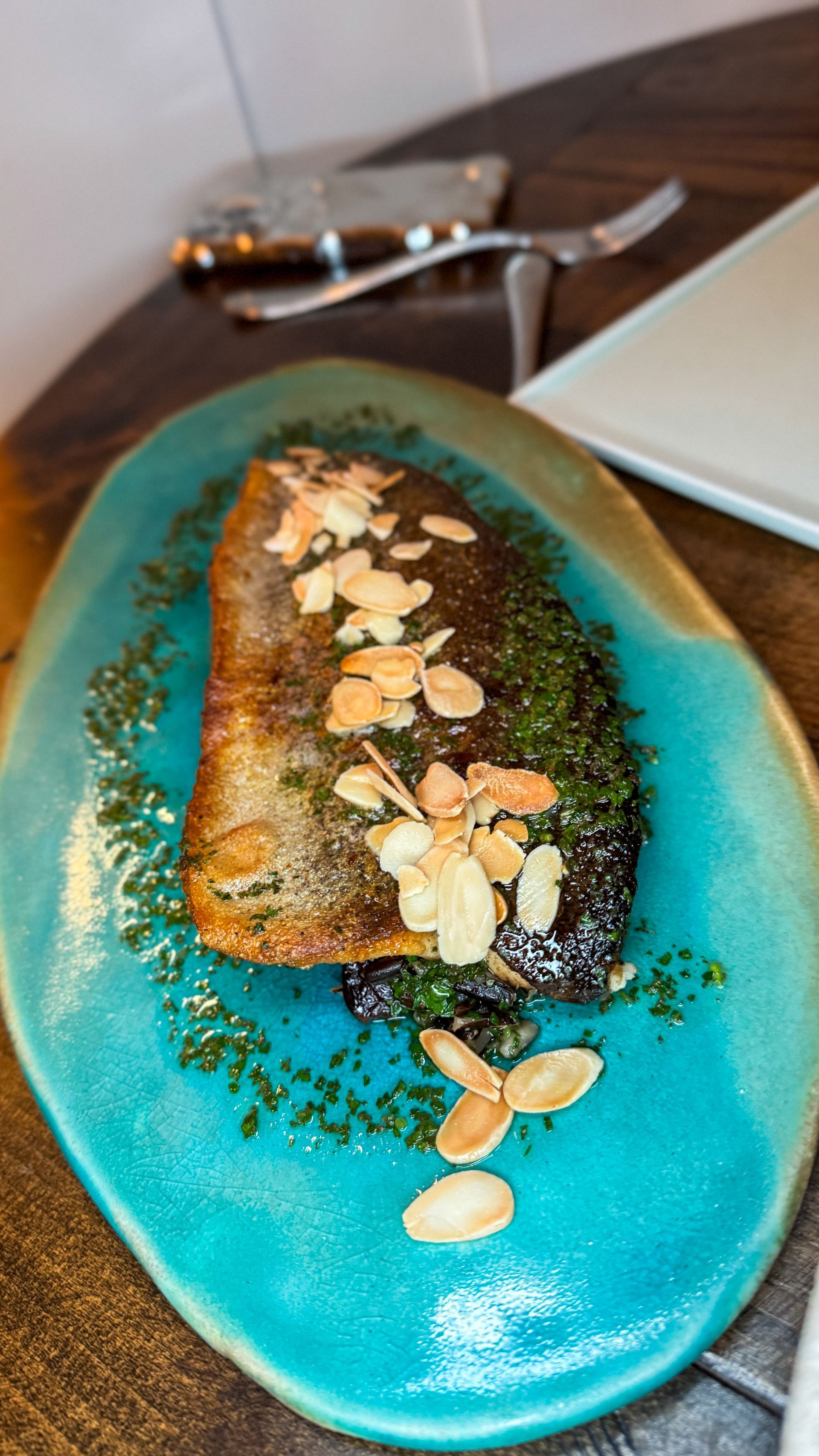 A pan-seared fish fillet topped with sliced almonds and herbs, served on a textured, turquoise-colored oval plate.