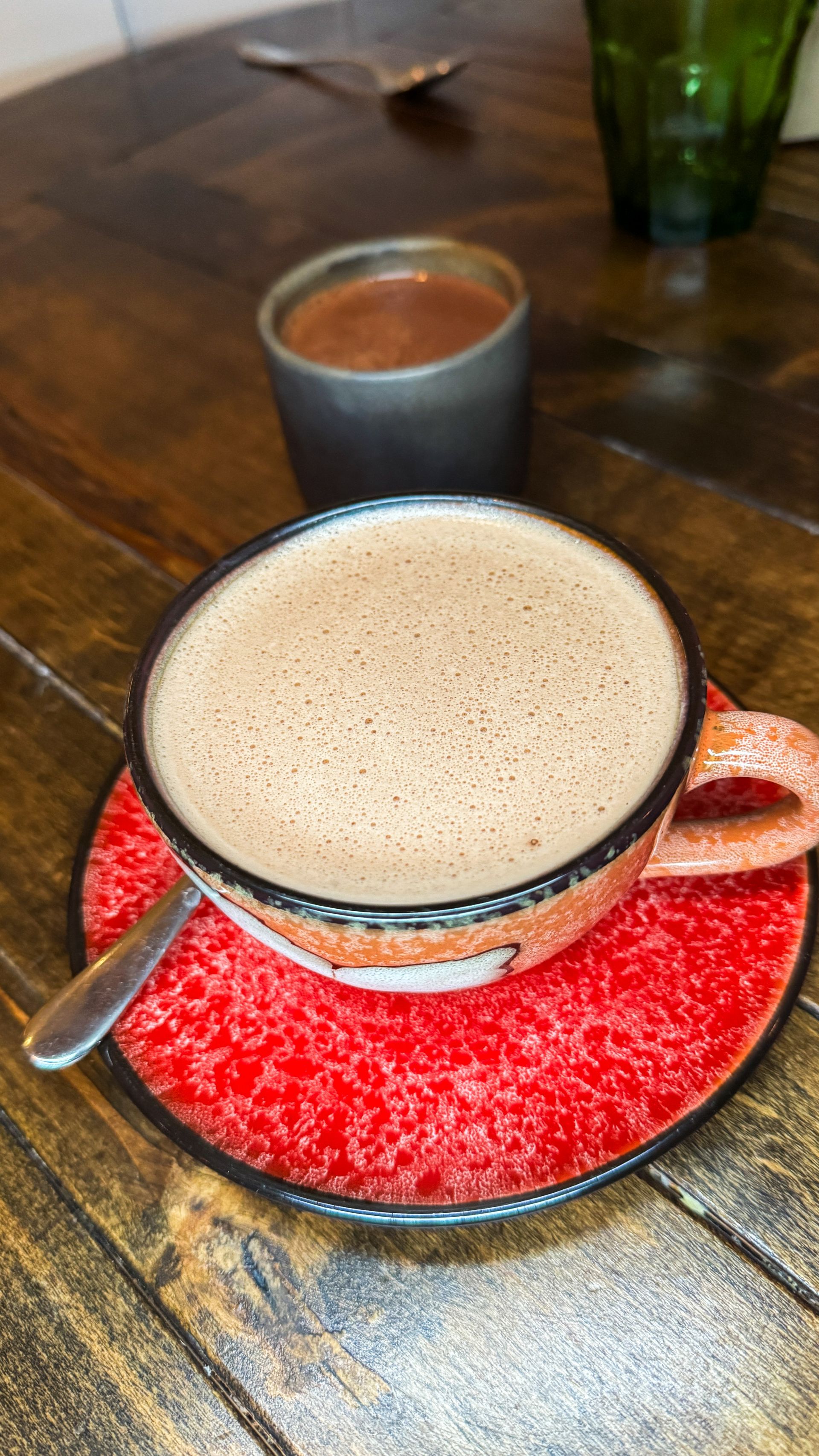 Cup of frothy coffee on a red saucer on a wooden table, with a dark mug behind it.