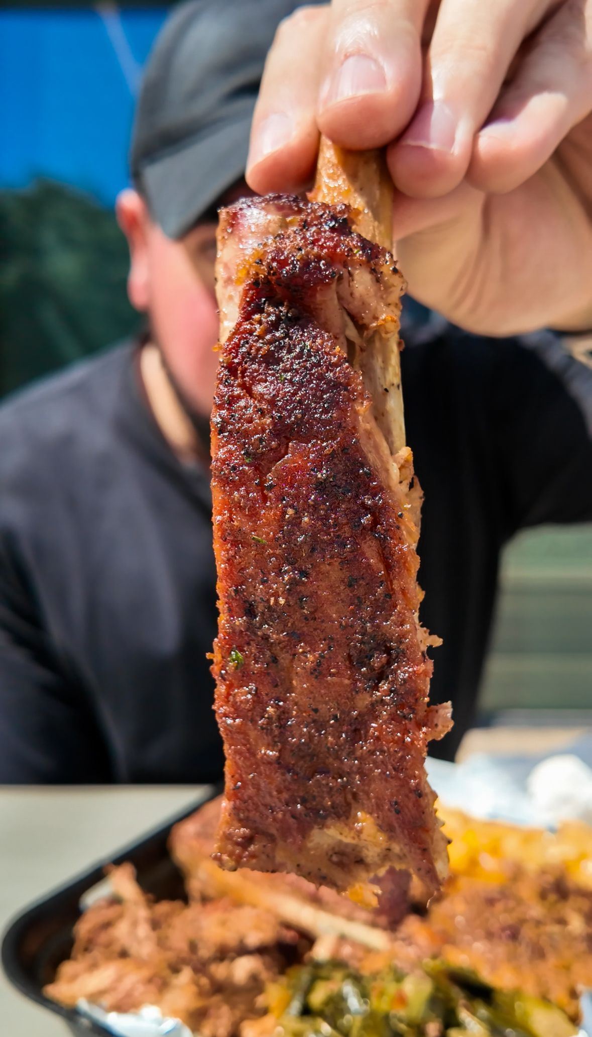 Hand holding a glazed, crispy rib over a plate of food, with a blurred person in the background