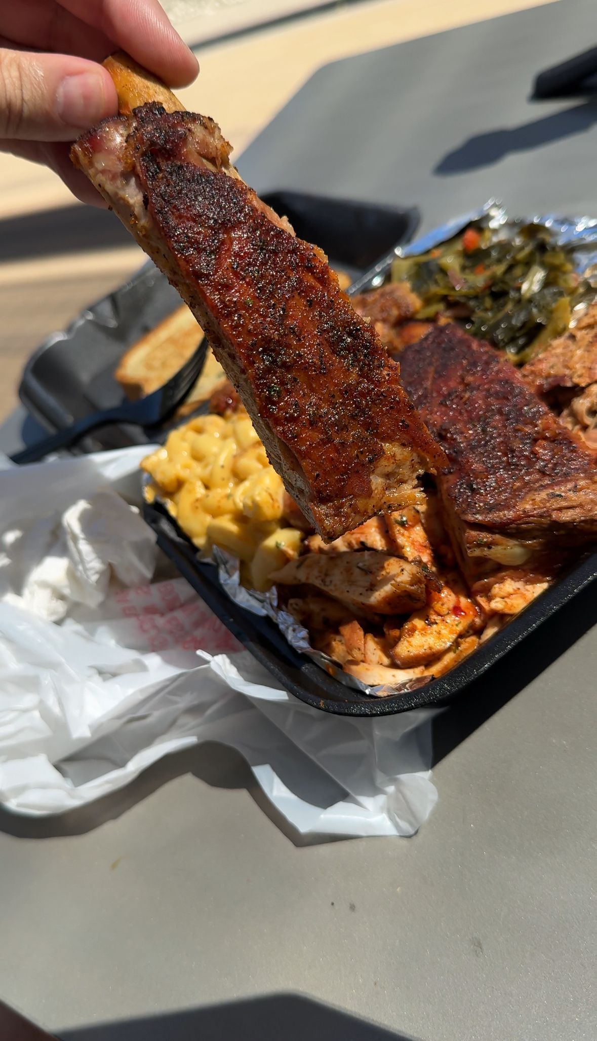 Hand holding a saucy smoked rib over a takeout tray with macaroni and collard greens.