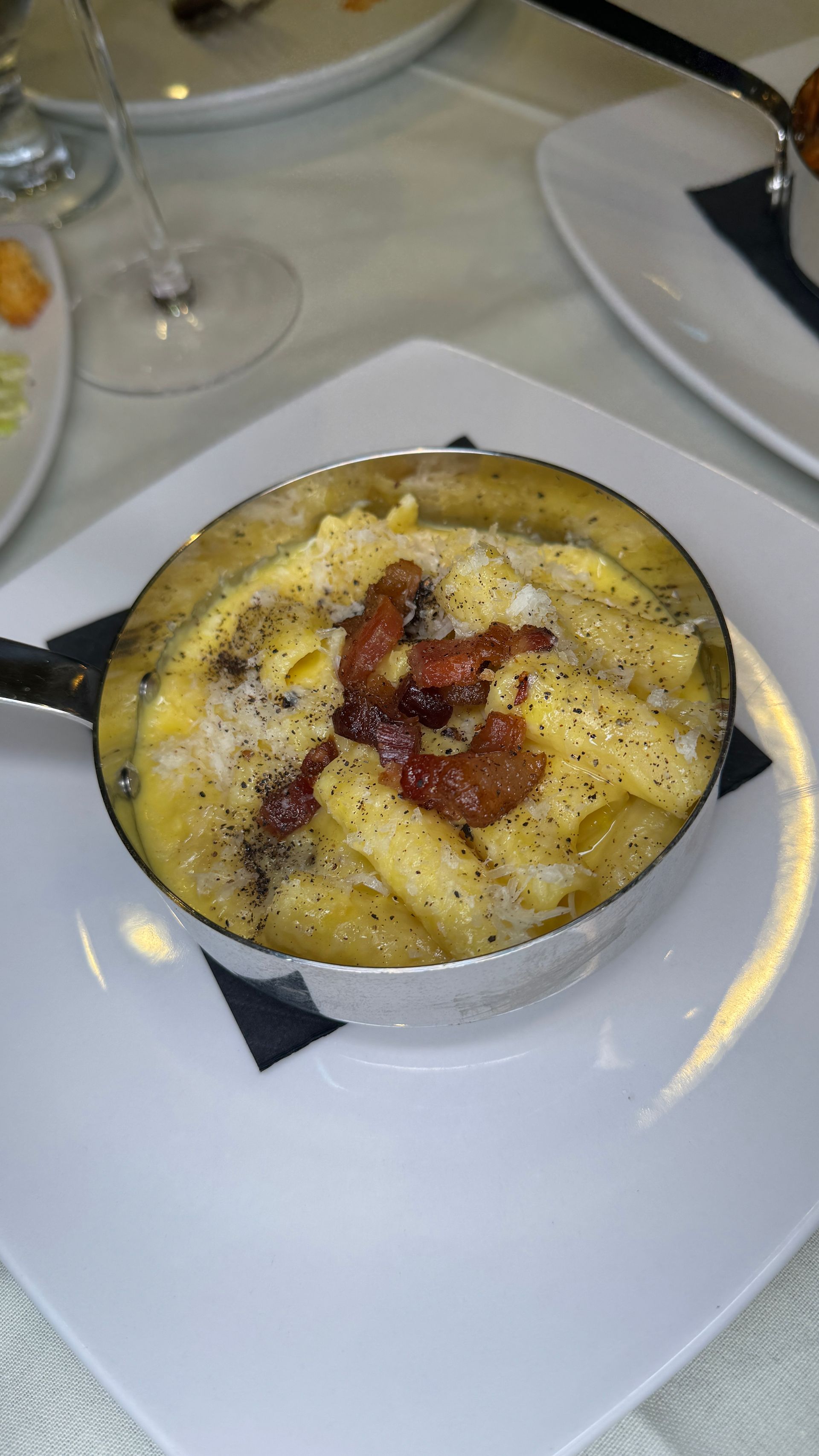 Macaroni and cheese in a metal dish, topped with bacon, on a white plate.