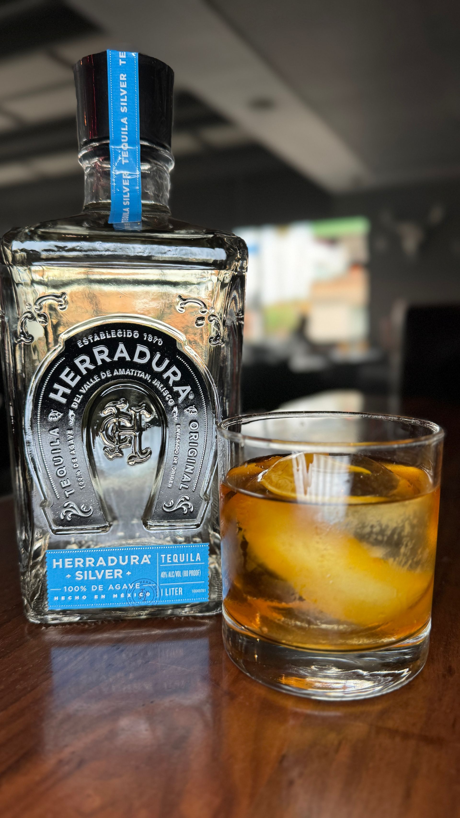 Bottle of Herradura tequila next to a glass with an amber-colored cocktail and ice sphere.